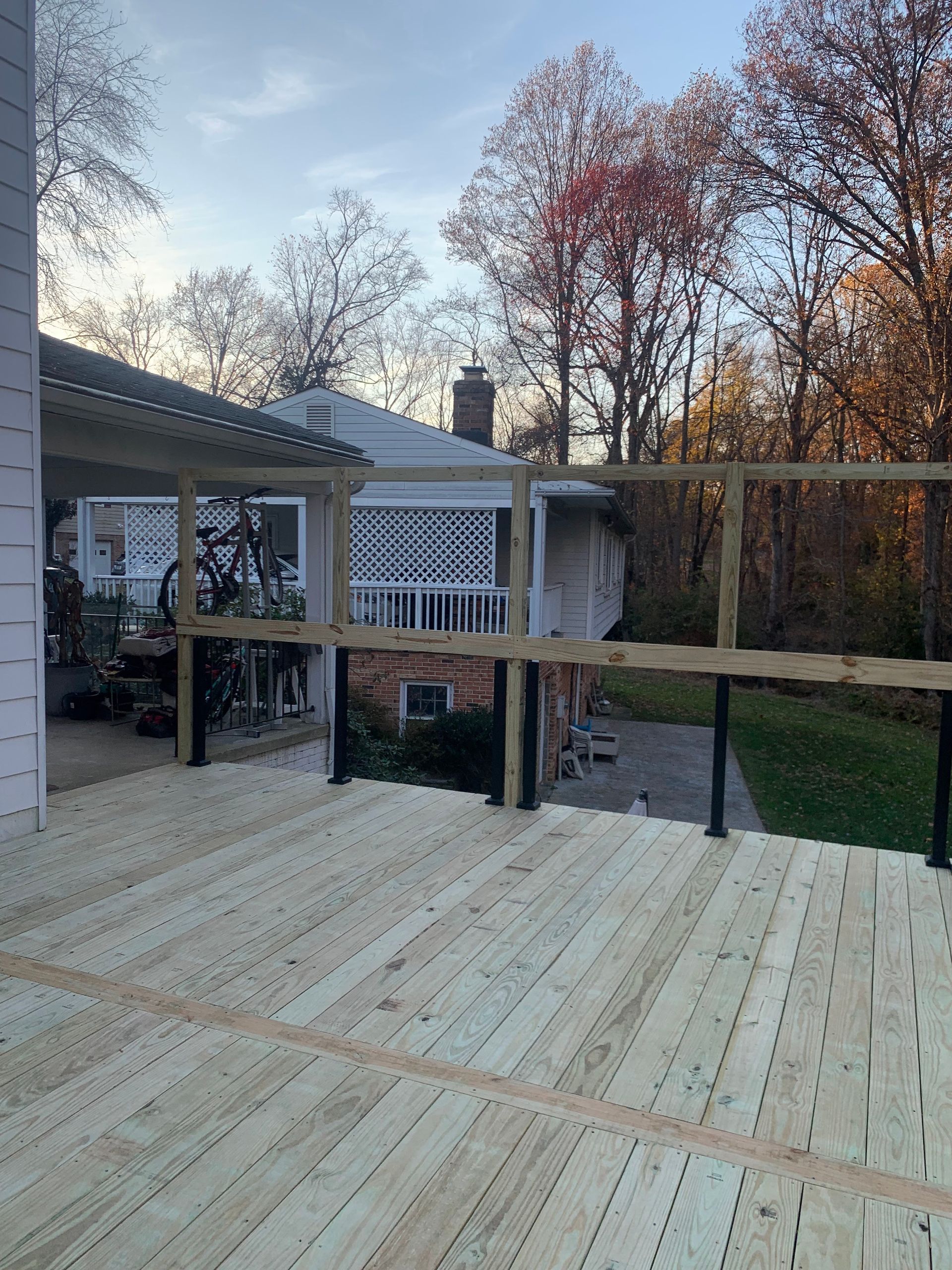Newly built wooden deck with railing, partially covered by a pergola. View of a house and trees in the background.
