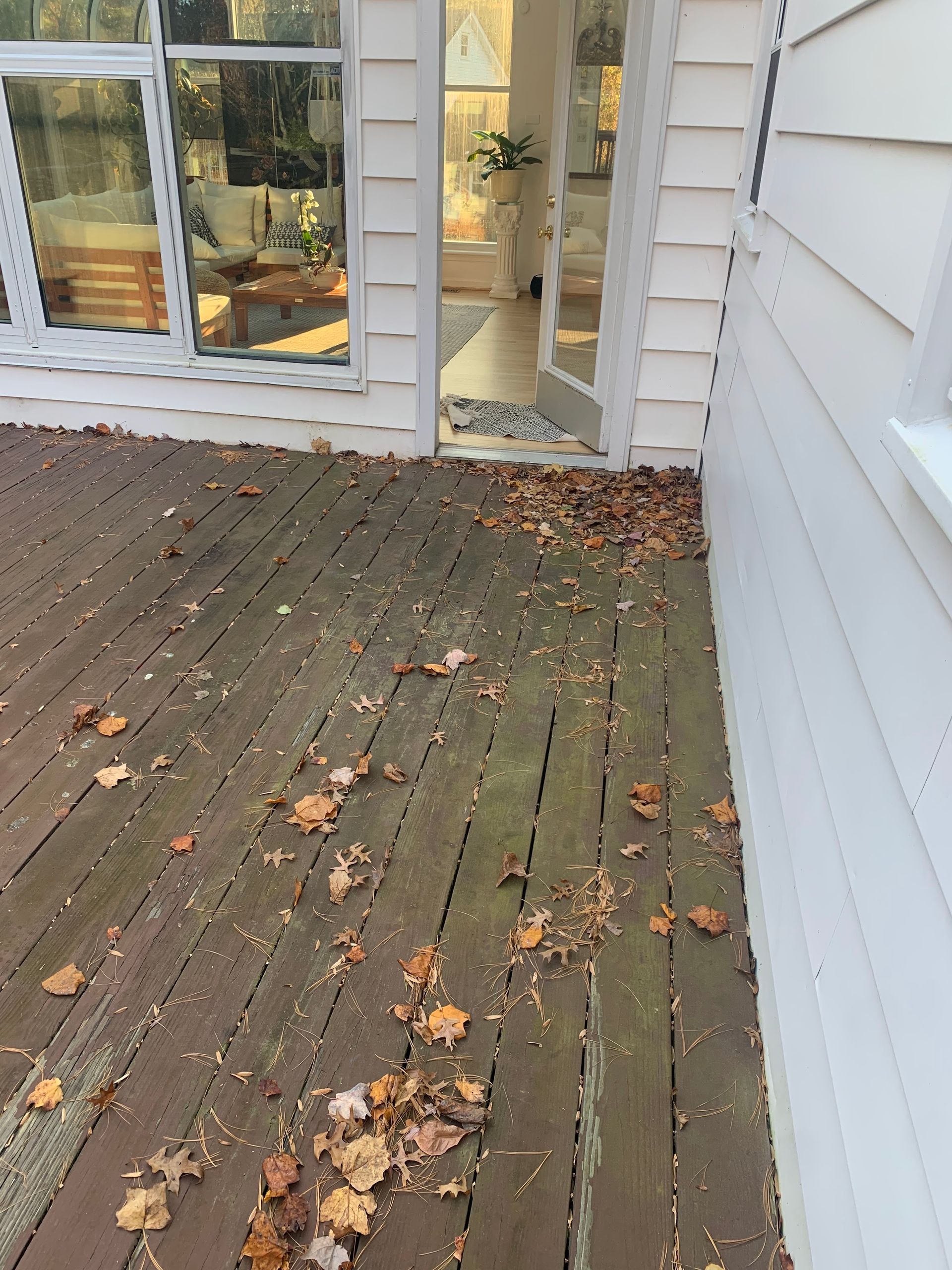 Wooden deck with scattered leaves, next to a white house with an open glass door.