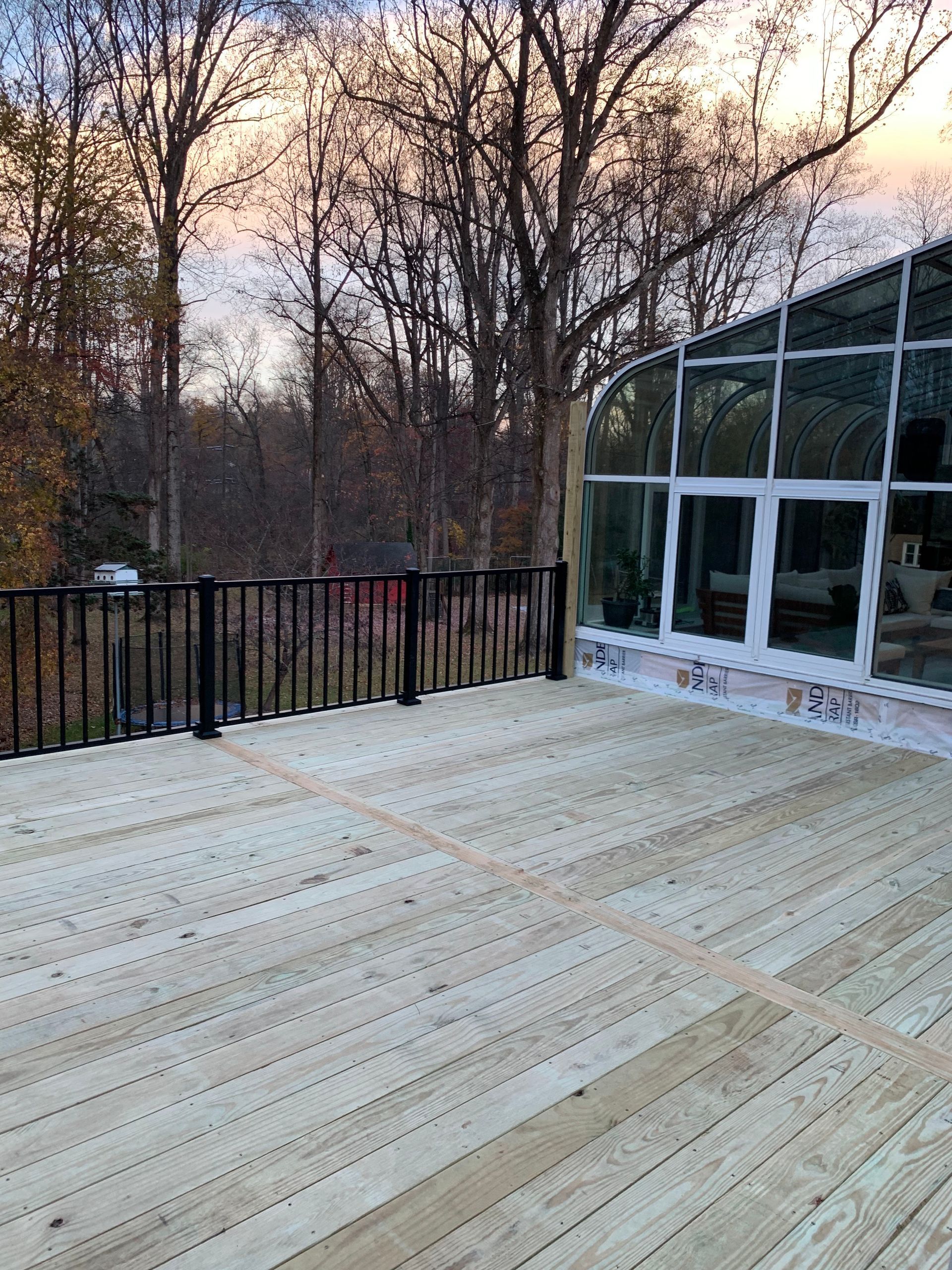 Wooden deck with black railing next to a glass-walled structure, surrounded by trees.
