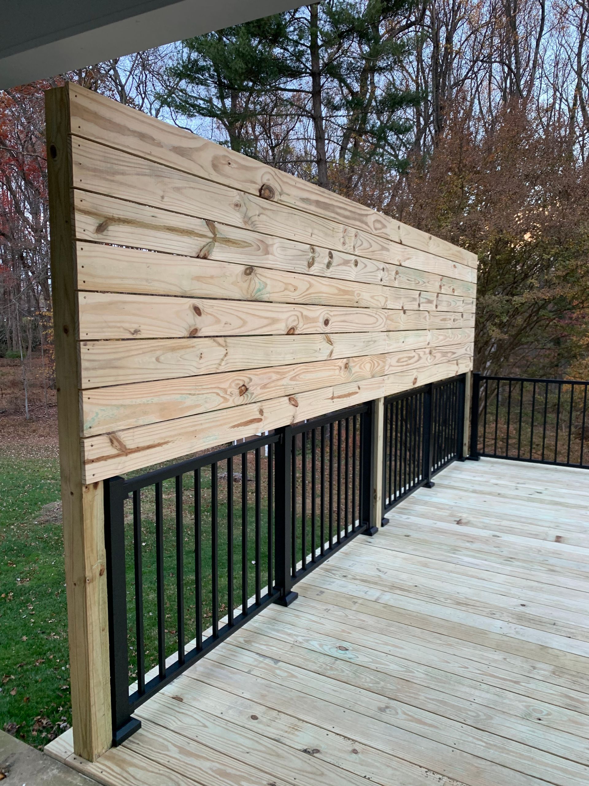 Wooden privacy screen on a deck with black railing. Wood deck and fall foliage in the background.