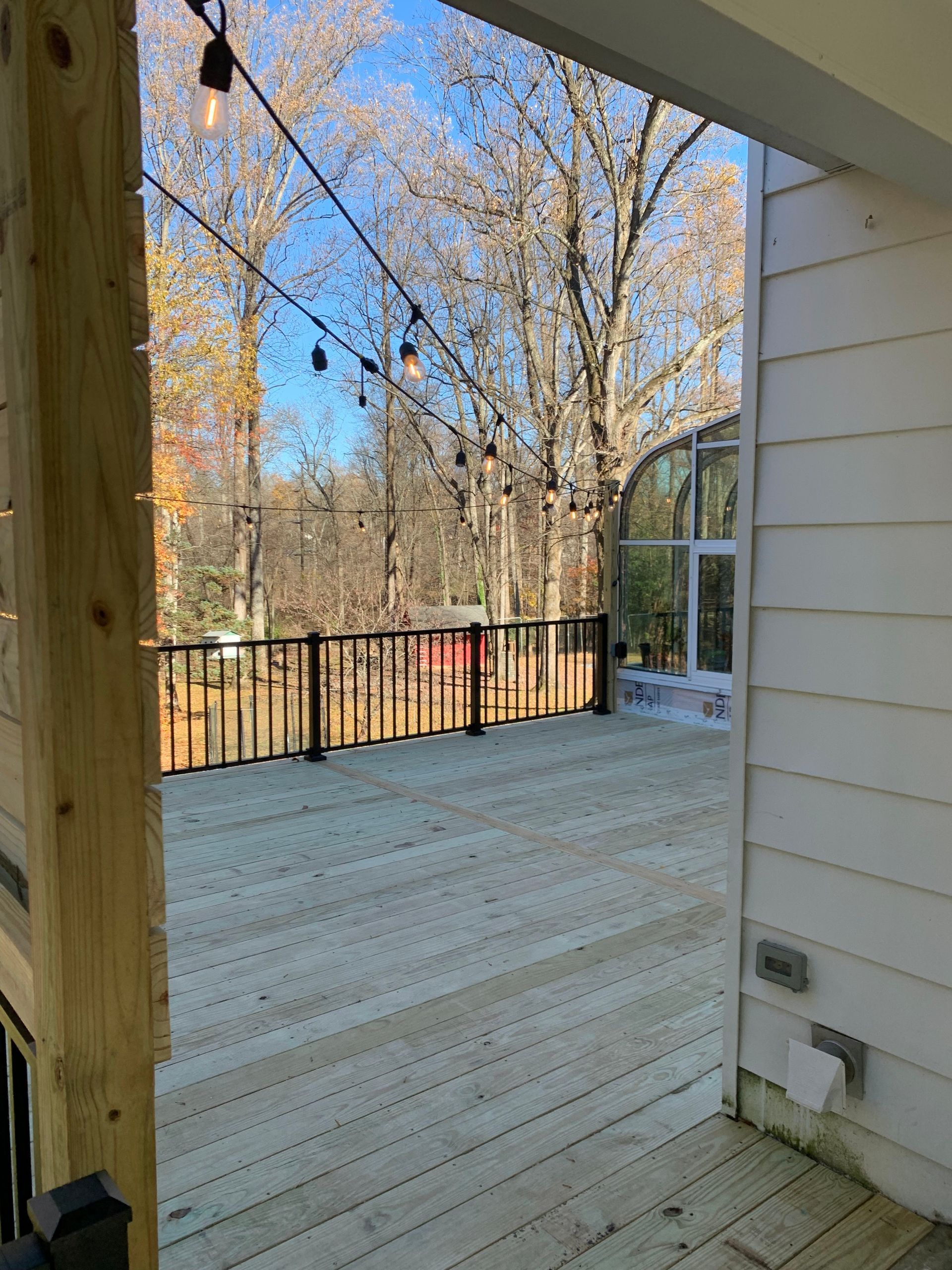 Wooden deck with black railing, string lights, and a view of trees under a blue sky.