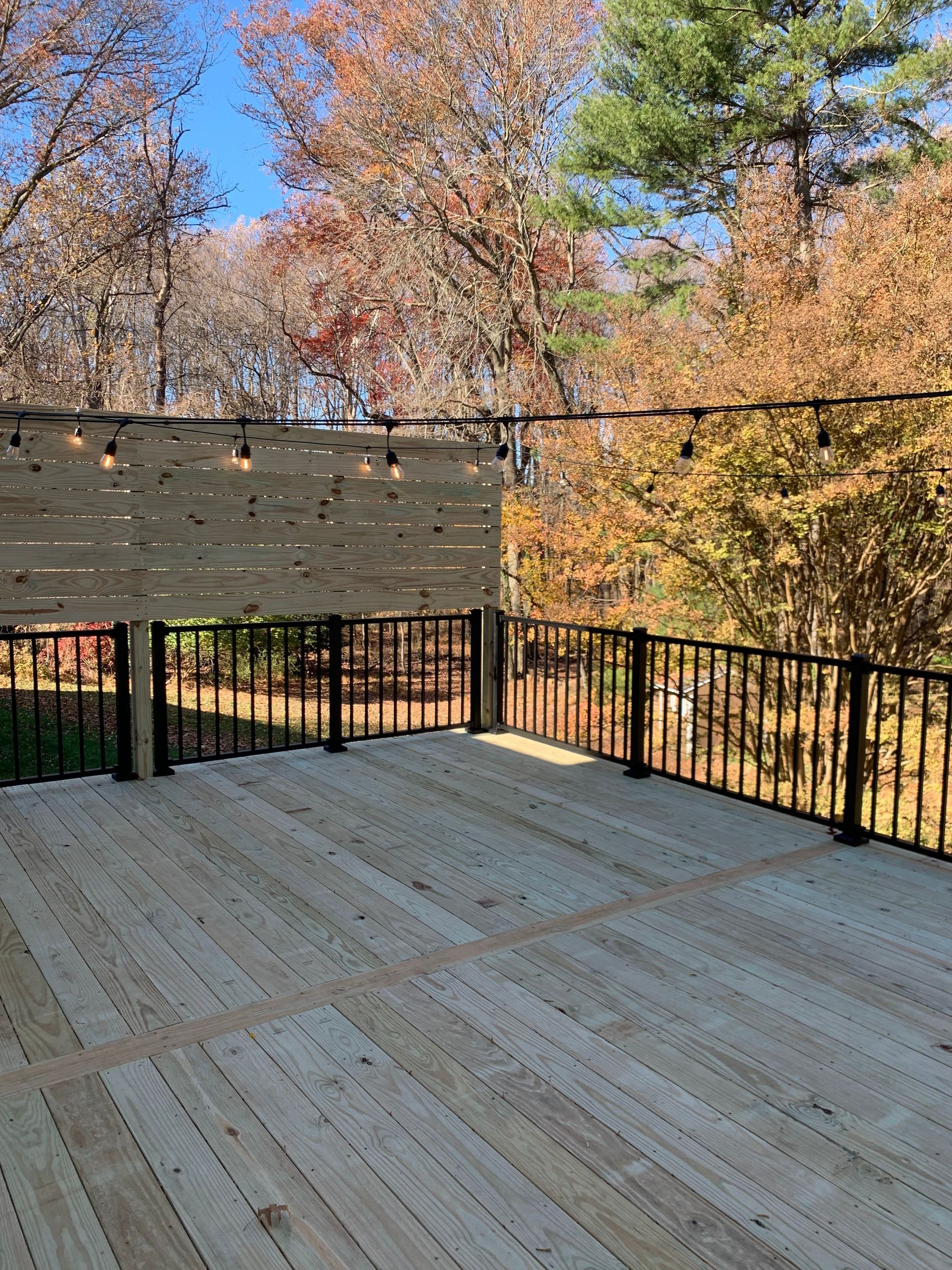 Wooden deck with black railings and light-strung wooden panel backdrop; trees with fall foliage in background.