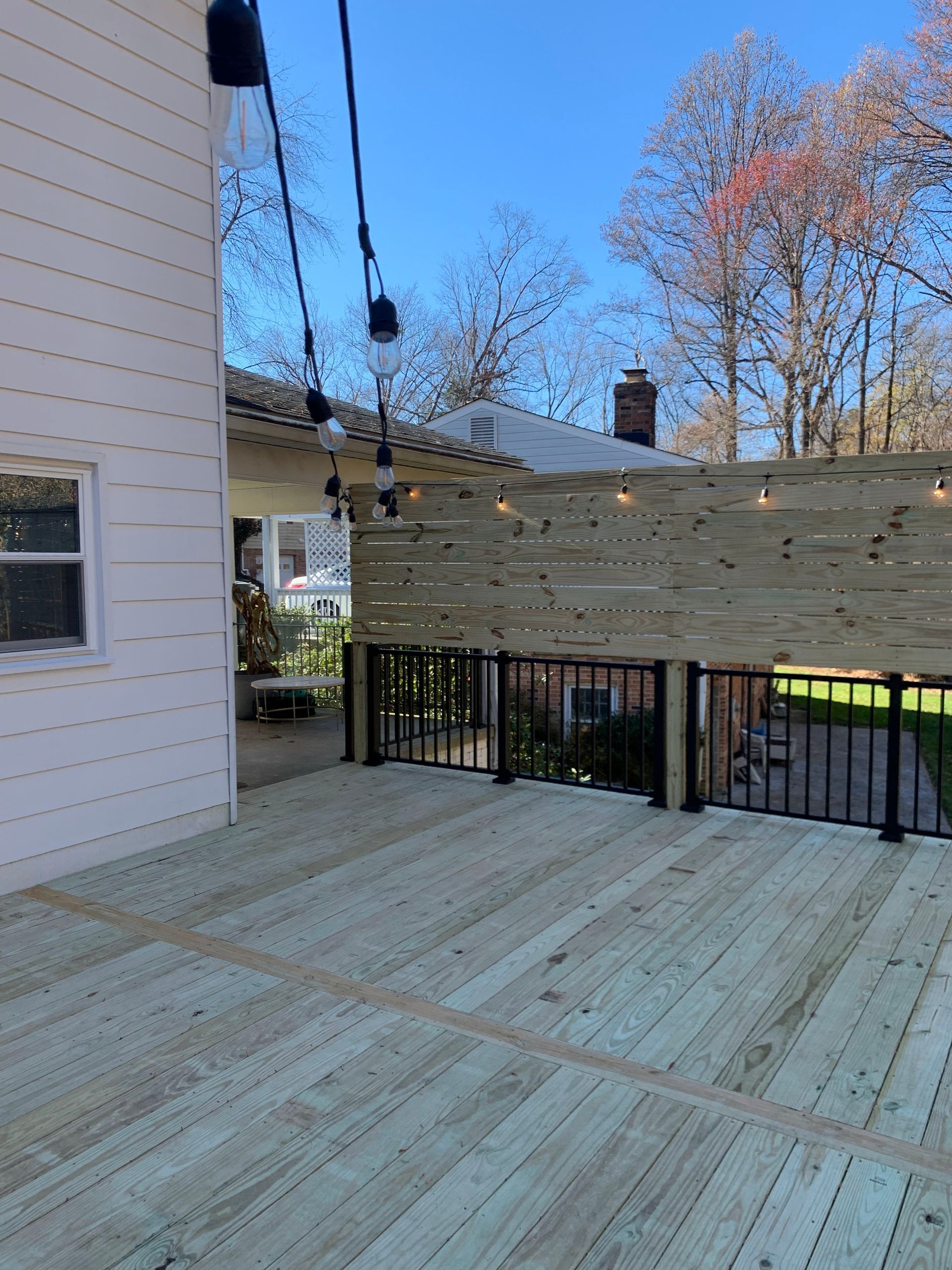 Wooden deck with string lights, fence, and a white house under a blue sky.