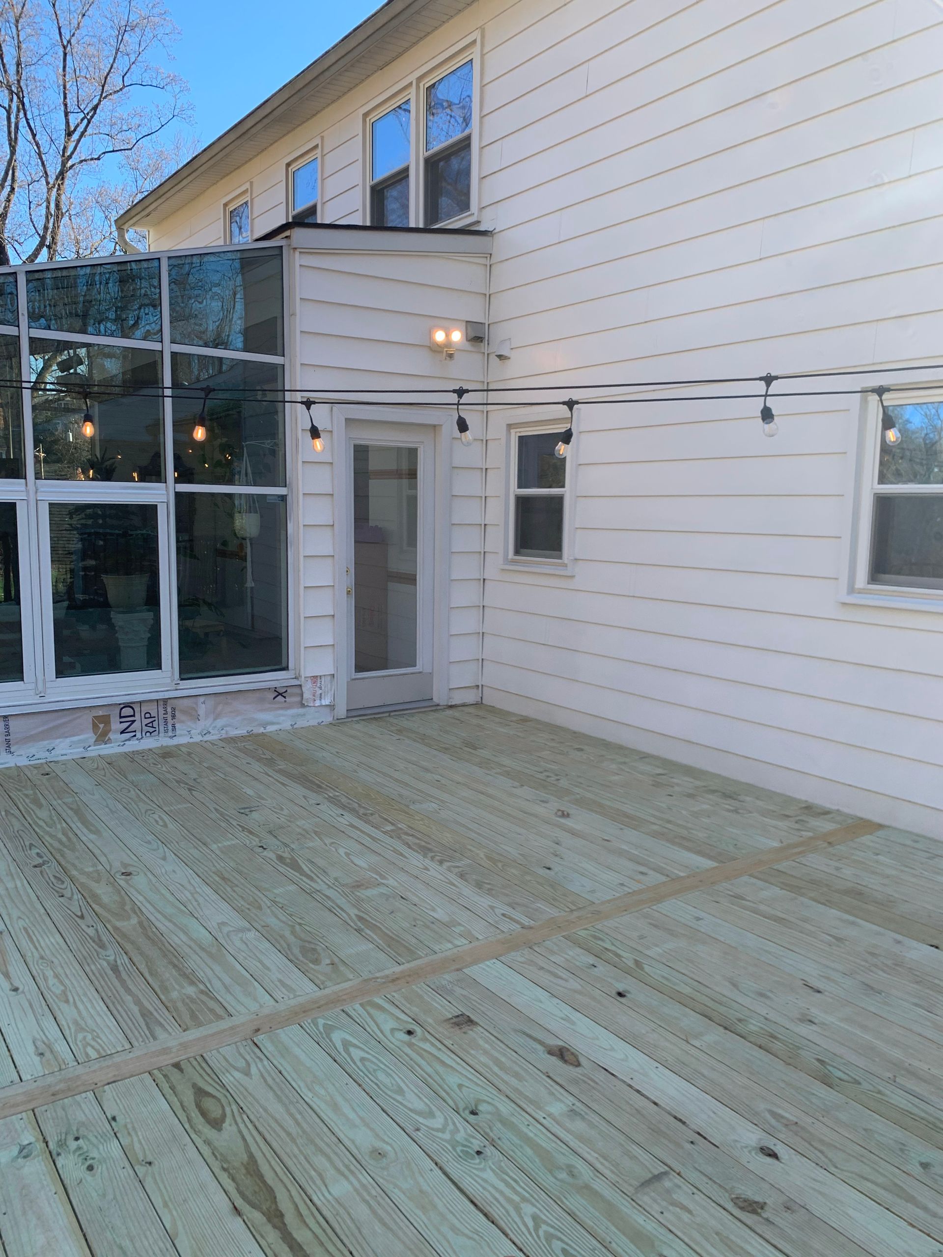 Newly built wooden deck attached to a white house with a glass-walled structure and string lights.