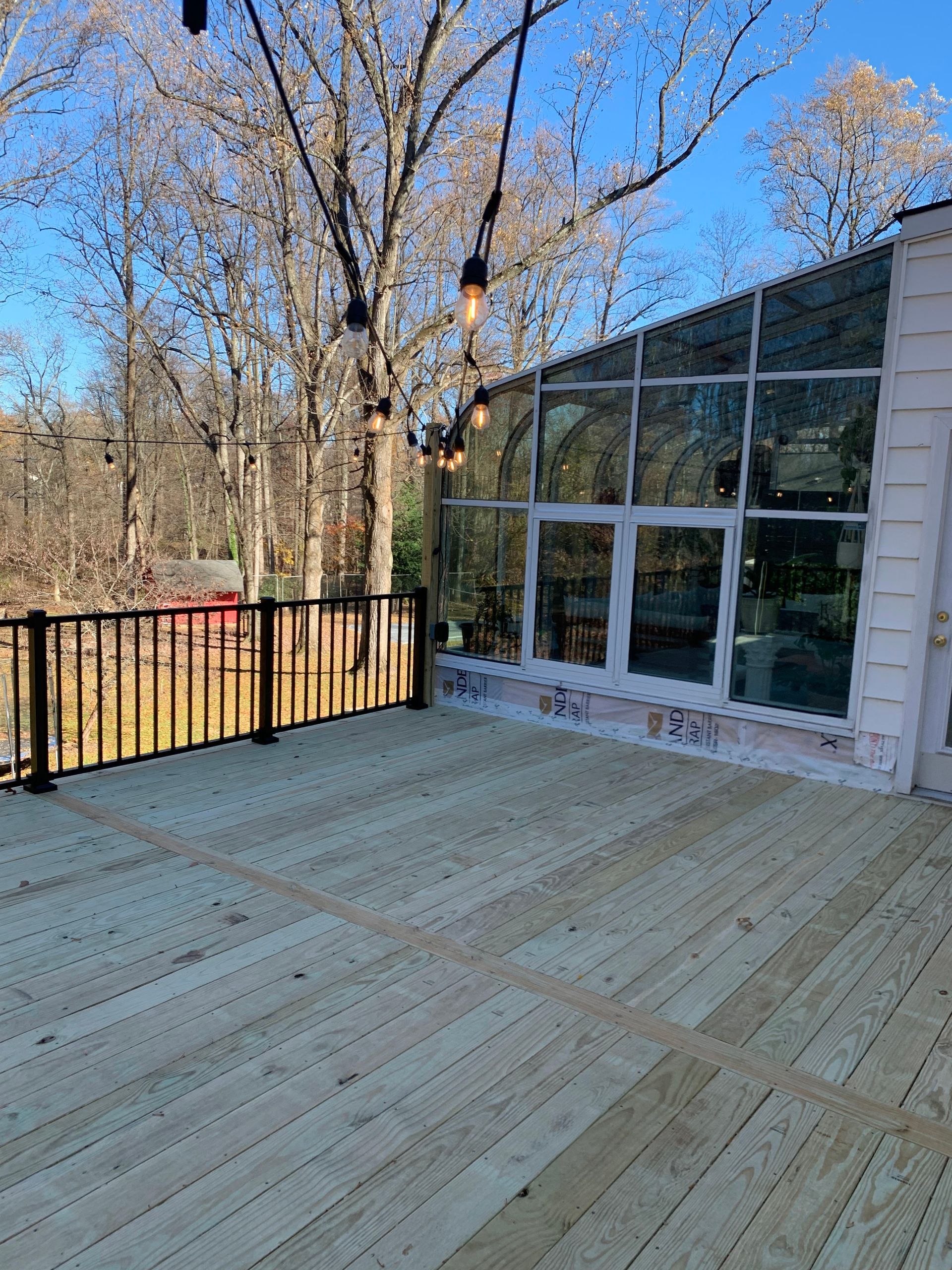 Wooden deck with black railing next to glass-walled sunroom and bare trees under a blue sky.