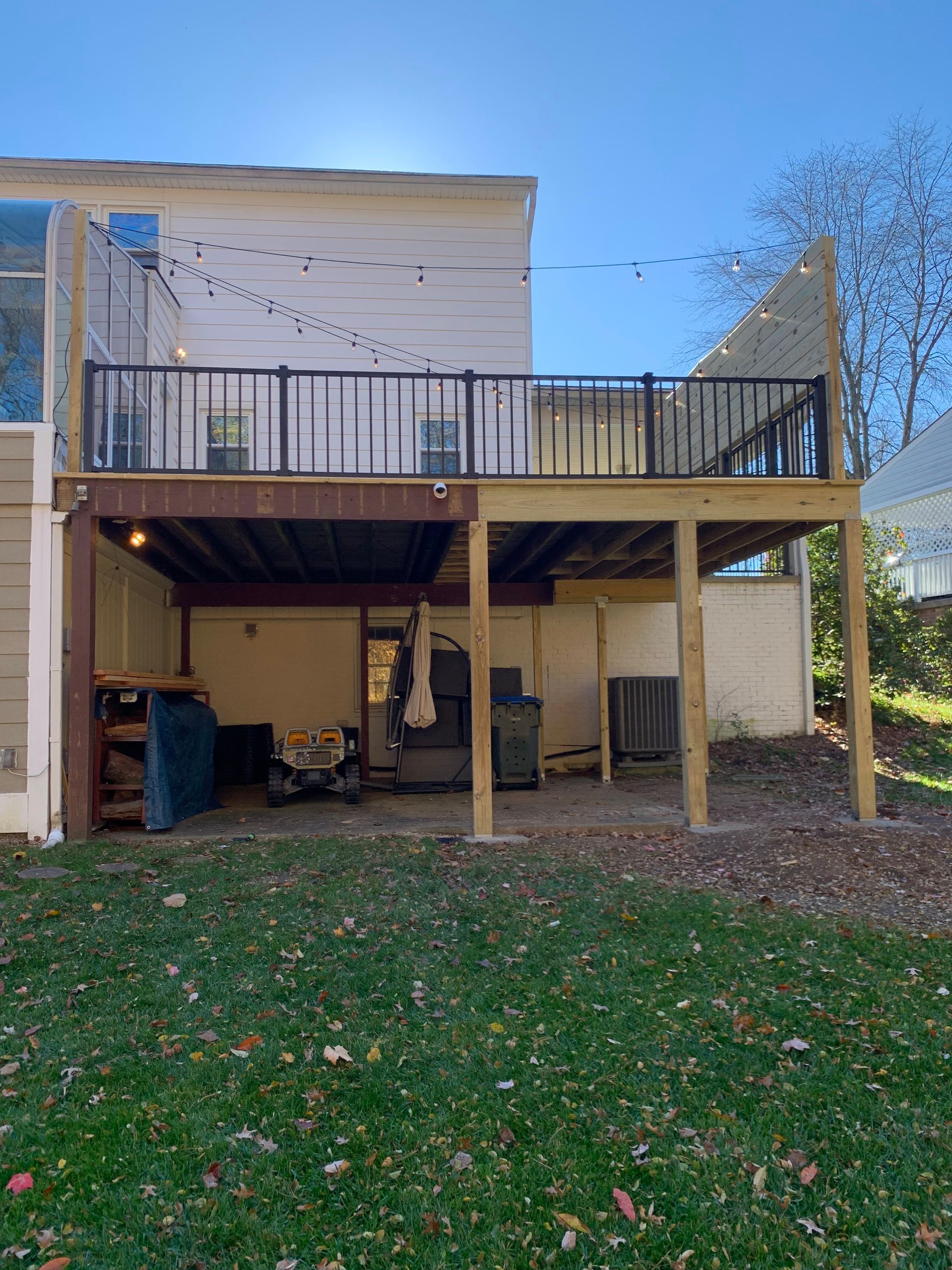 Two-story house with a wooden deck. The deck has a black railing. Underneath is a storage area with tools.