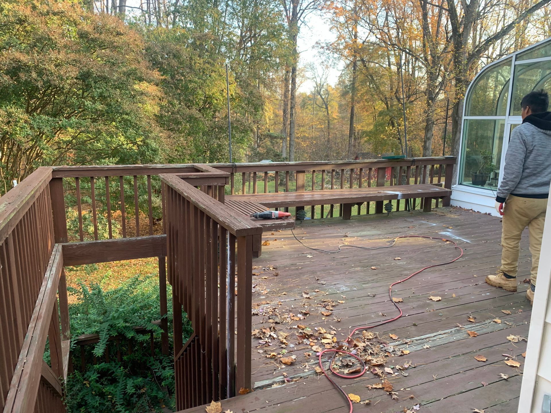 Person on wooden deck, fall foliage visible. Deck has benches and access stairs, leaves scattered.