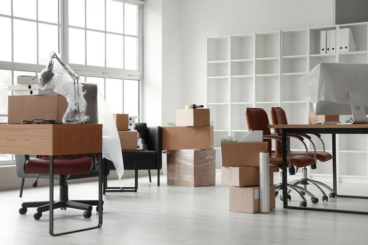 Office space being moved out: furniture, boxes, and a desk covered in plastic wrap sit in a bright room.