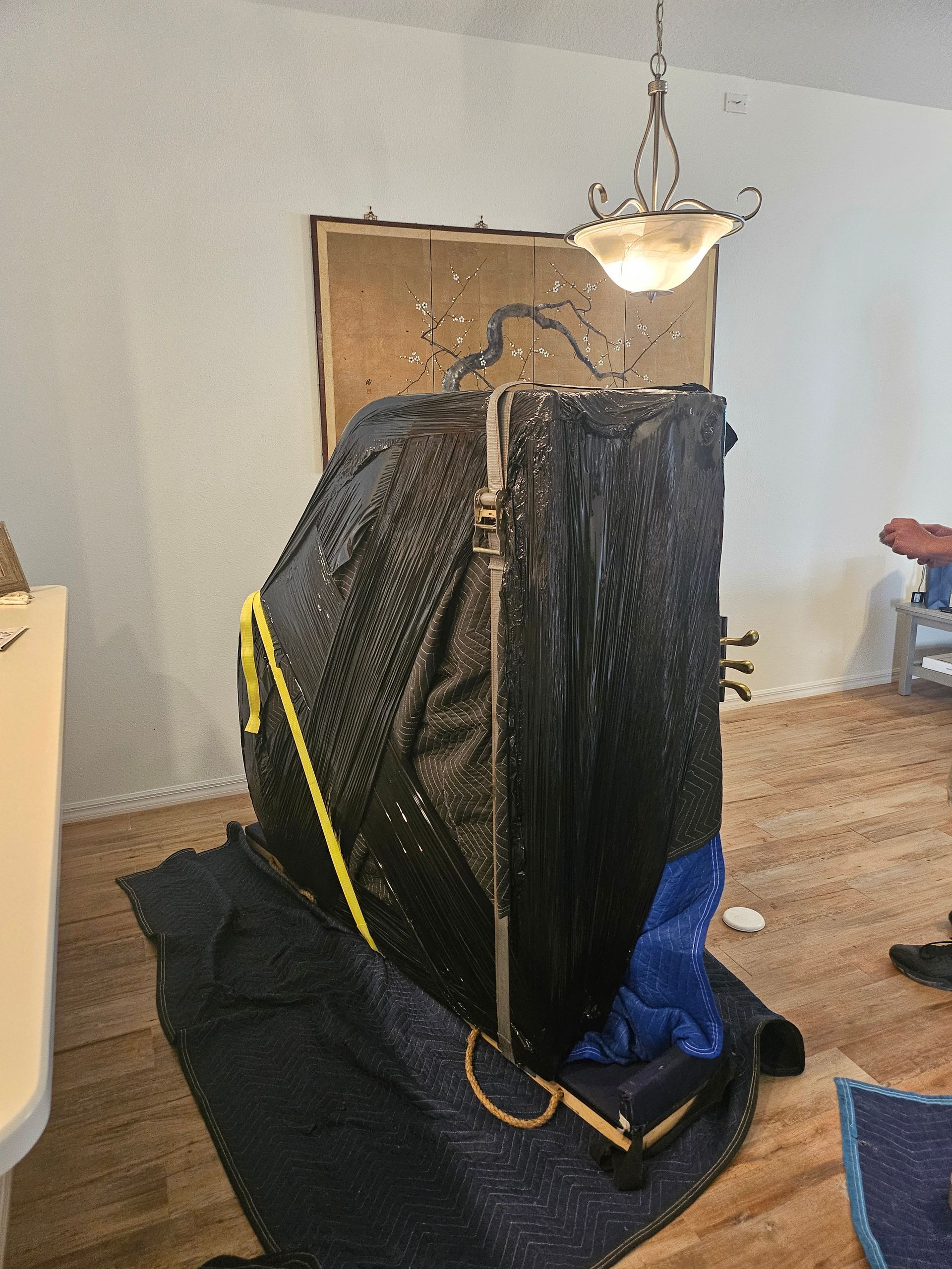 A large, covered object, likely a piano, is being moved indoors. It's wrapped in protective black material and sits on a rug, with moving supplies around.