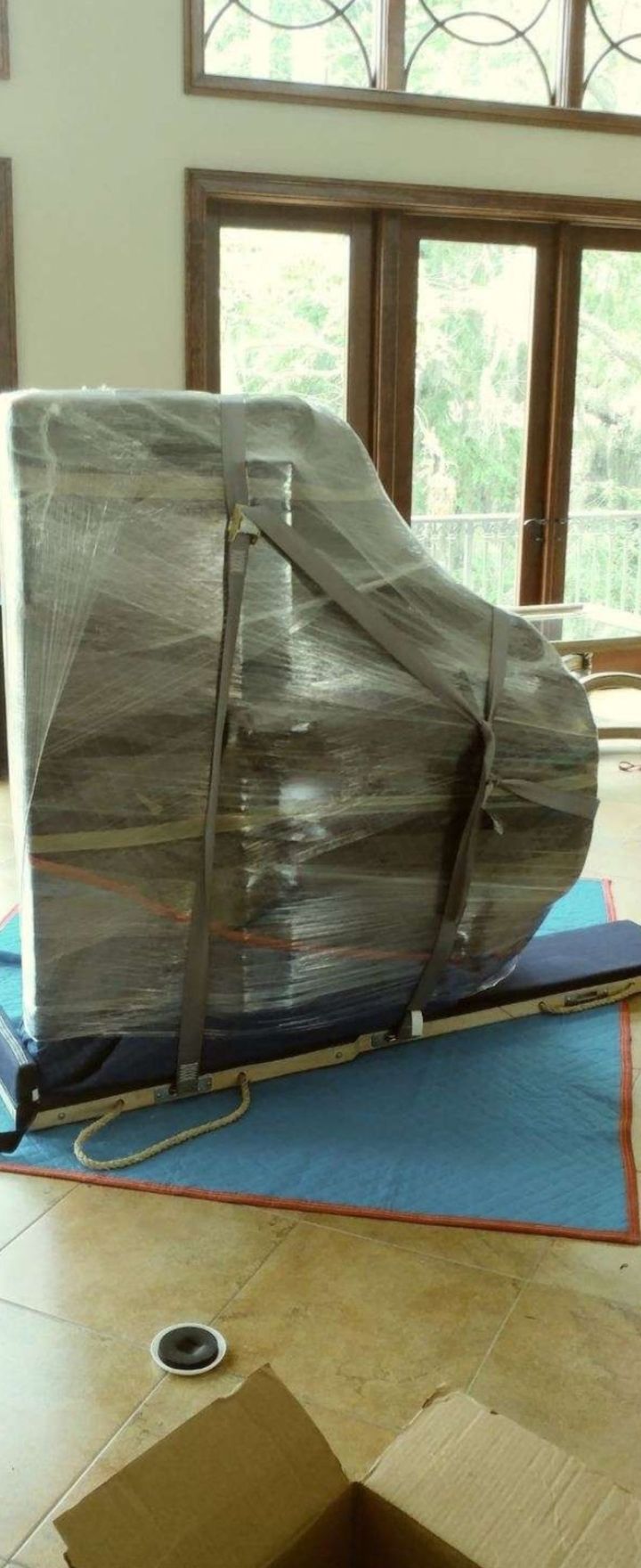 A grand piano wrapped in plastic and secured with straps, possibly being moved, inside a room with large windows and wooden floors.