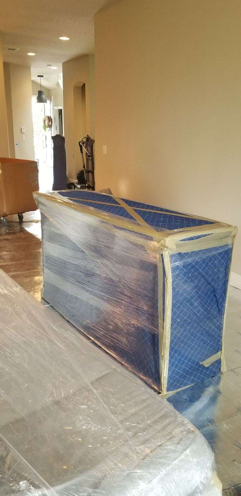 A piece of furniture wrapped for moving, covered in blue padding and clear plastic wrap, sits in a beige-walled room. Another wrapped piece is on the left.