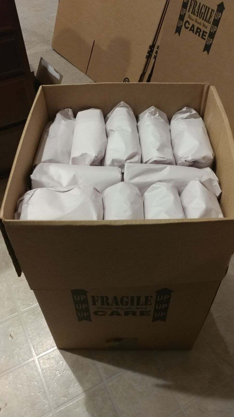 Cardboard box filled with wrapped items, likely glasses or mugs, labeled