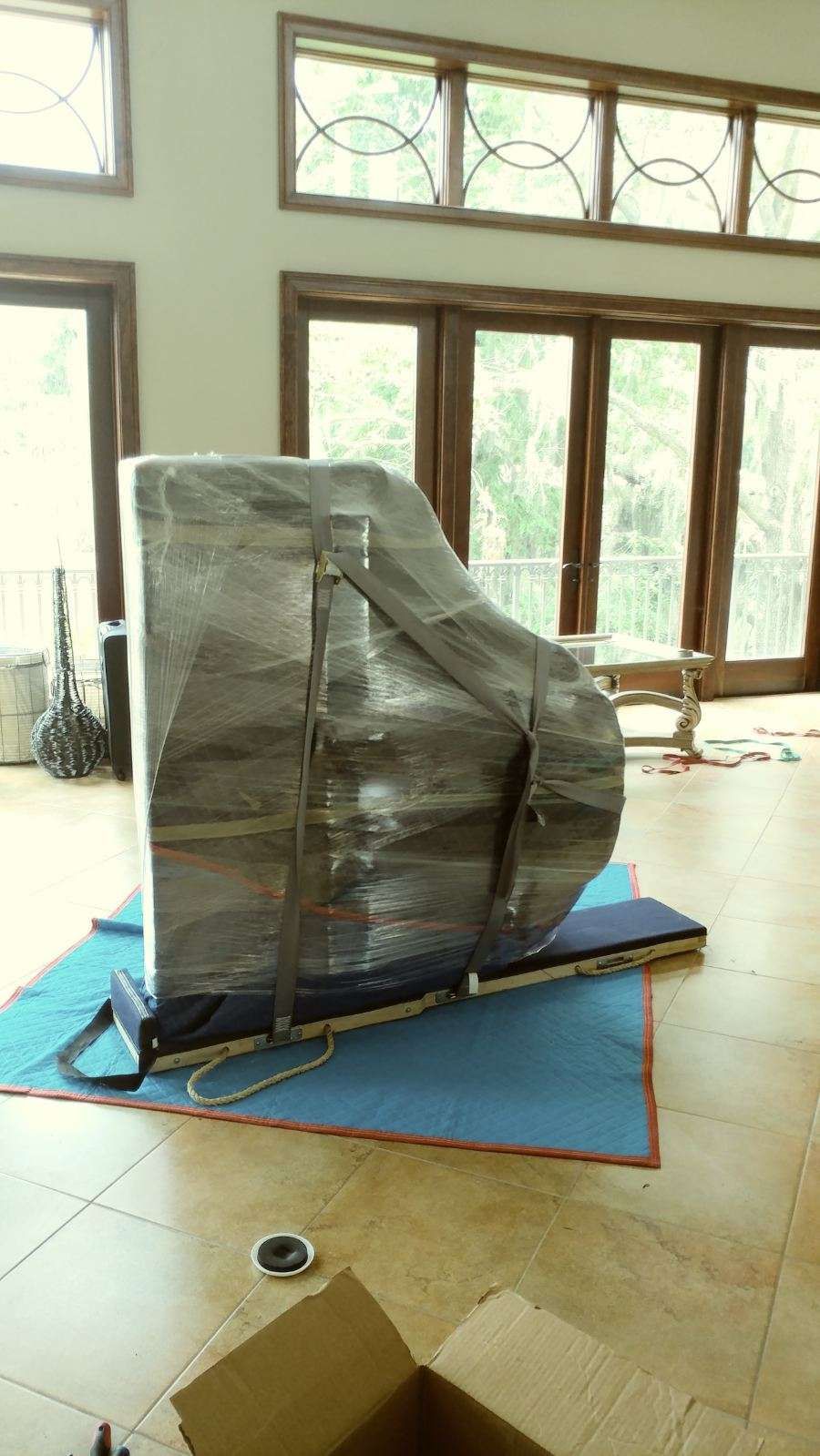 A piano wrapped in plastic and secured with straps, prepared for moving, inside a room with large windows.