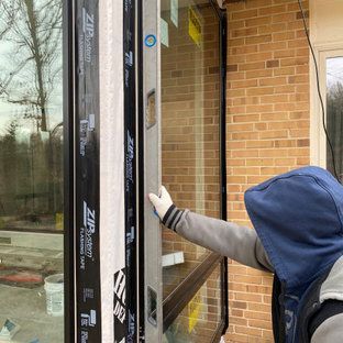 A person is standing in front of a brick building holding a window.