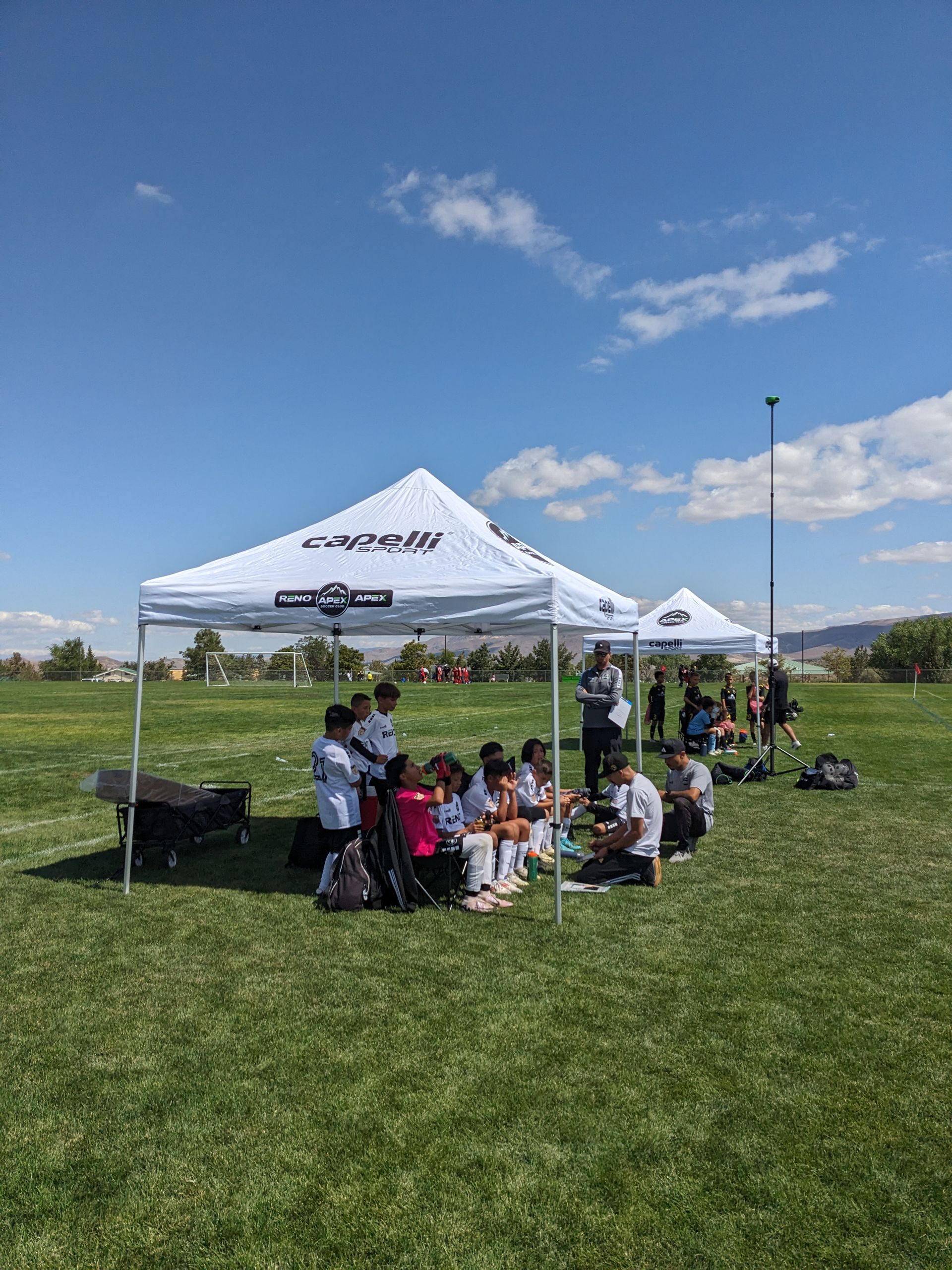 A group of players and coaches are sitting under tents in a grassy field at half time of a soccer game.