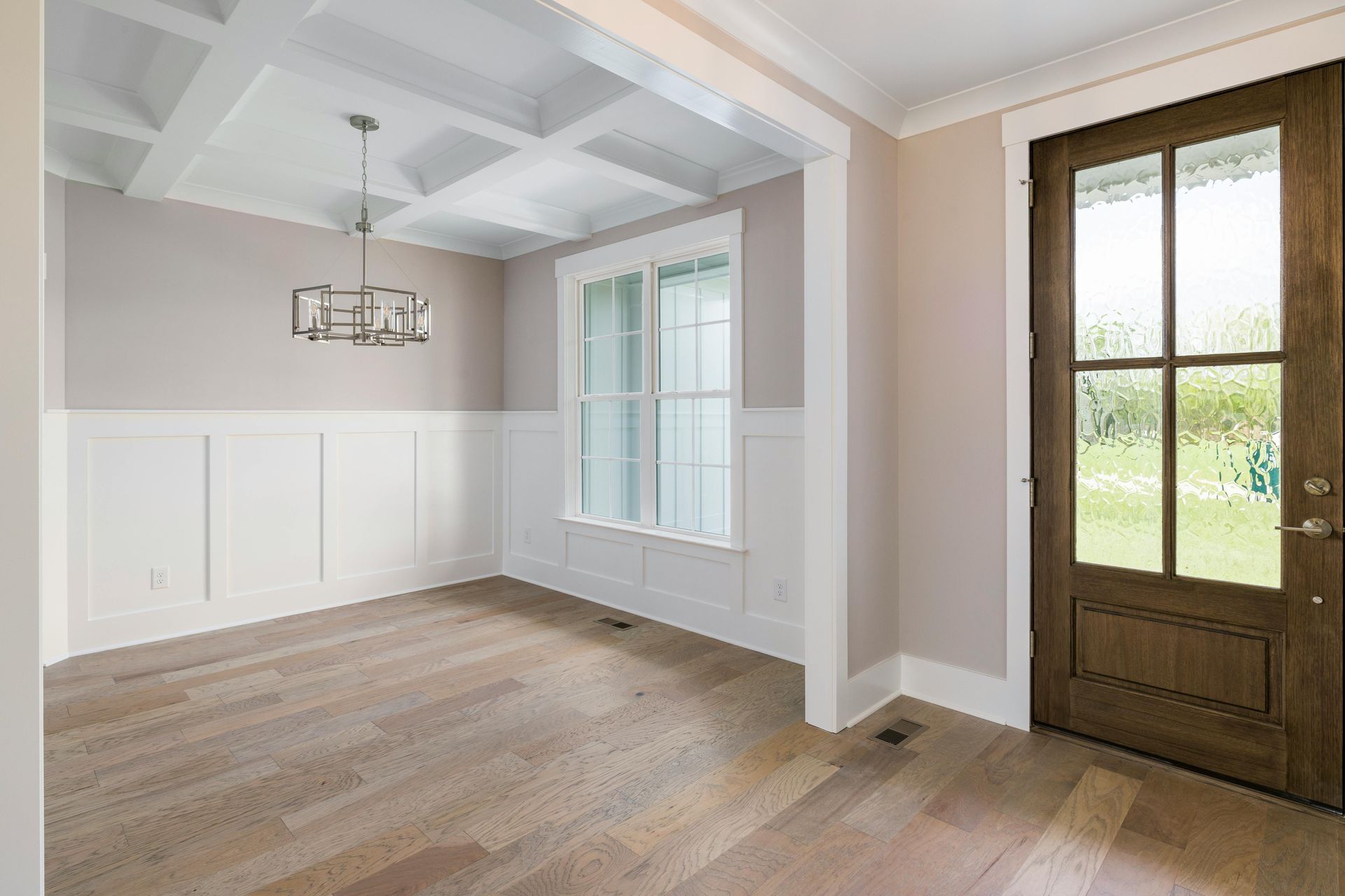 Empty room with wooden floors, wainscoting, and a coffered ceiling. A window and front door are visible.