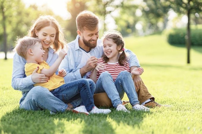 Family sitting on grass, laughing and smiling in a park on a sunny day.