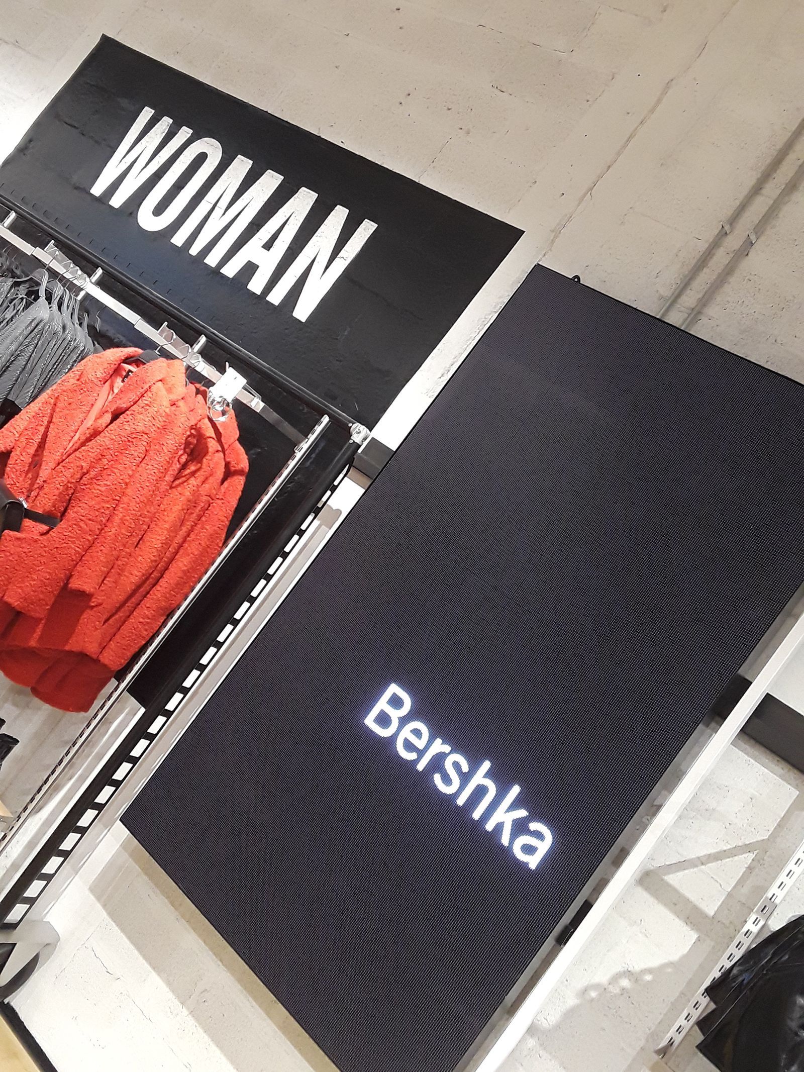 Exhibición de tienda de ropa Bershka, cartelería negra: “MUJER”, “Bershka”, suéter rojo visible.