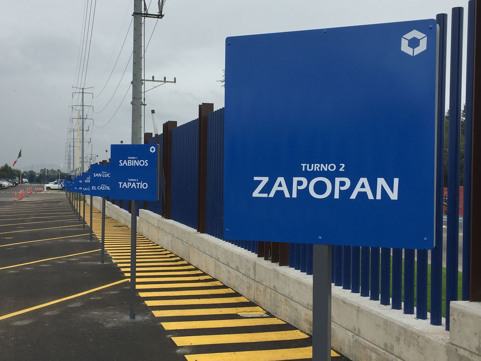 Señales azules con texto blanco indican dirección a Zapopan. Están junto a un muro con una acera gris y líneas amarillas pintadas sobre el asfalto.