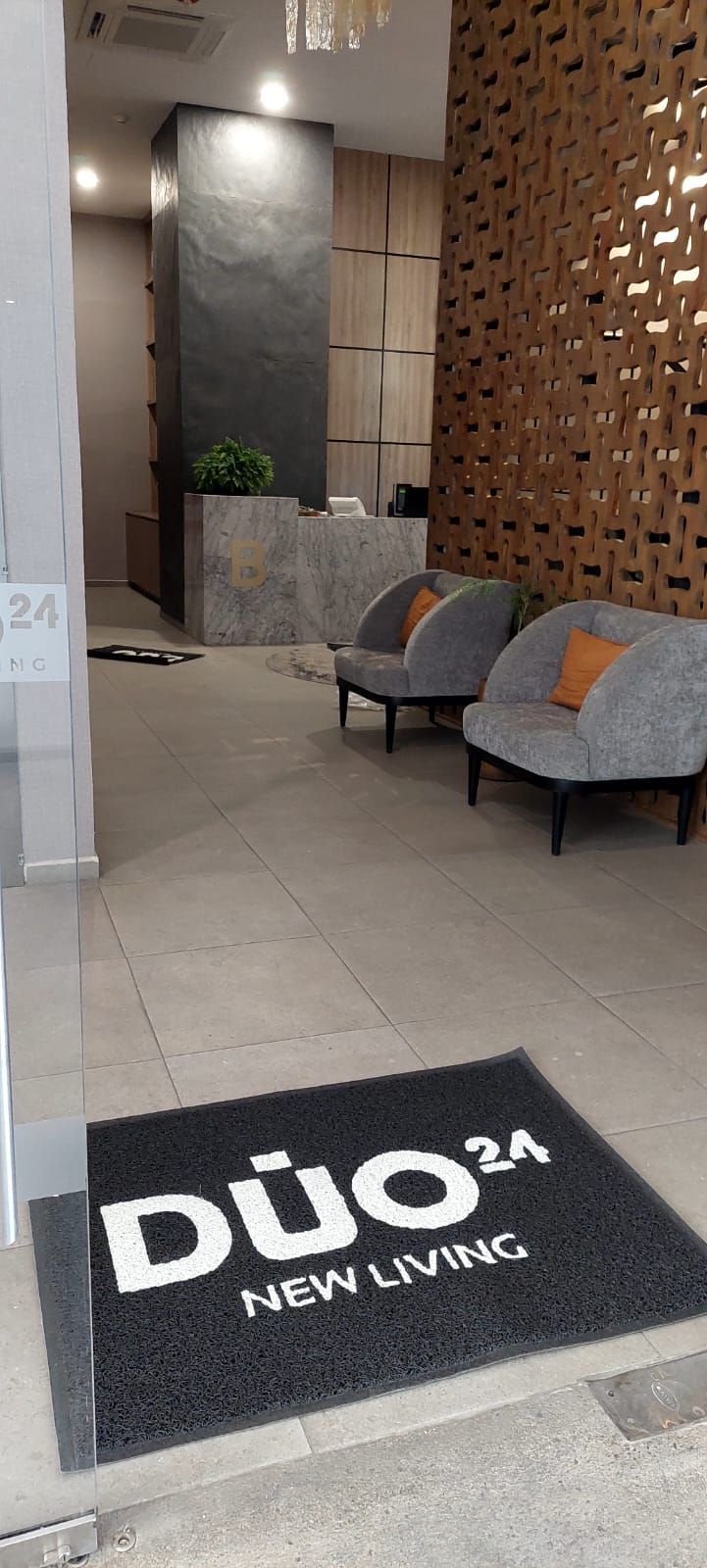 Entrada al edificio “Duo 24 New Living” con felpudo, sillas y diseño moderno.