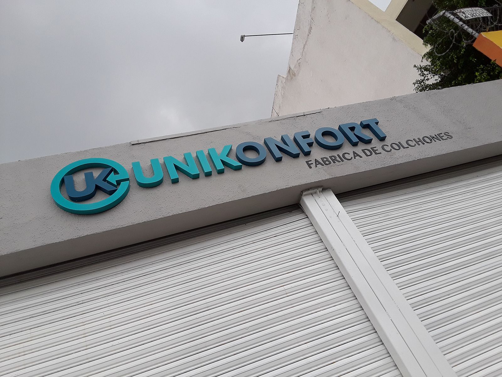 Cartel de Unikonfort, una fábrica de colchones, con letras azules y verde azulado sobre un edificio gris.