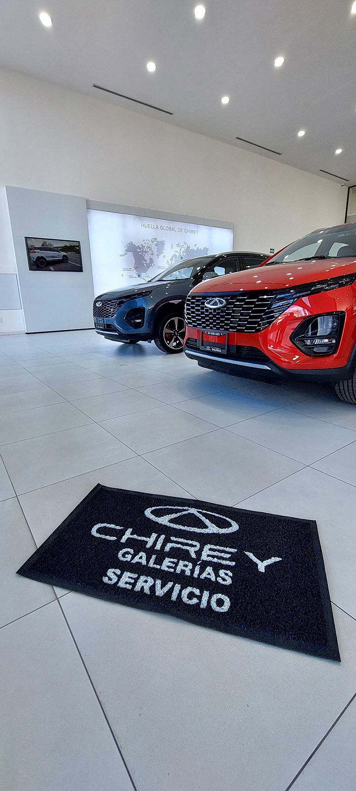 Coches aparcados en una sala de exposición con una alfombra con el logo de Chery. Coches rojos y azules sobre un suelo de baldosas bajo luces brillantes.