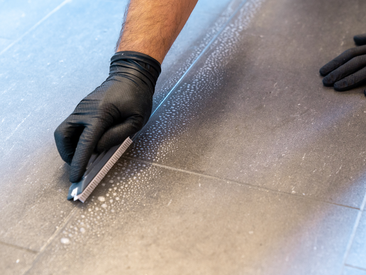 Gloved hands scrubbing grout on a gray tiled floor with a rectangular tool.