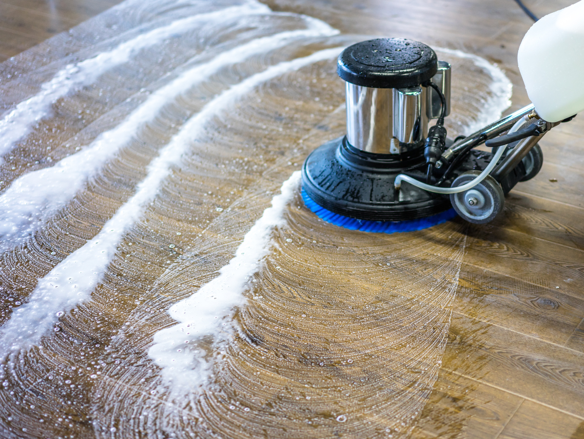Floor cleaning machine scrubbing a wood floor with white soapy solution.