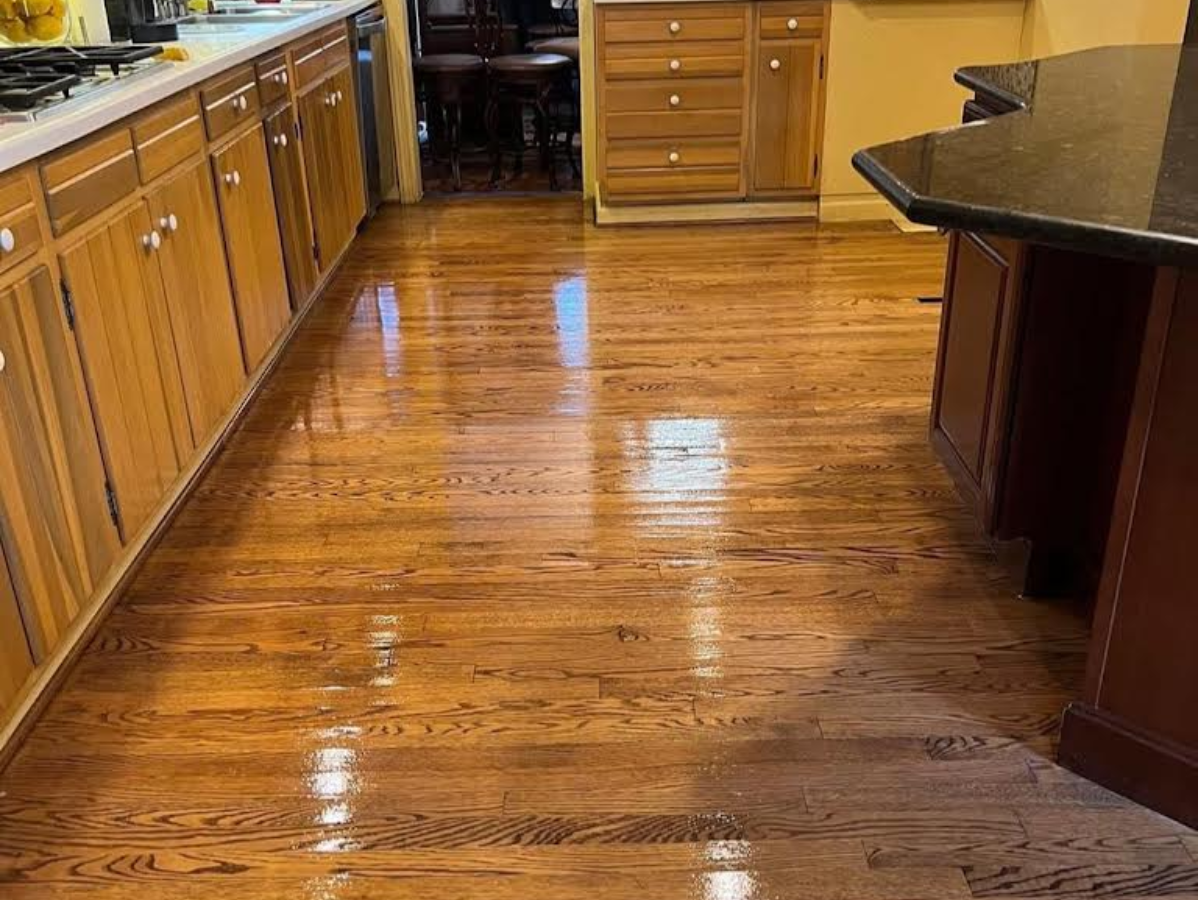 Hardwood kitchen floor with shiny finish, reflecting light. Brown cabinets on left and island on right.