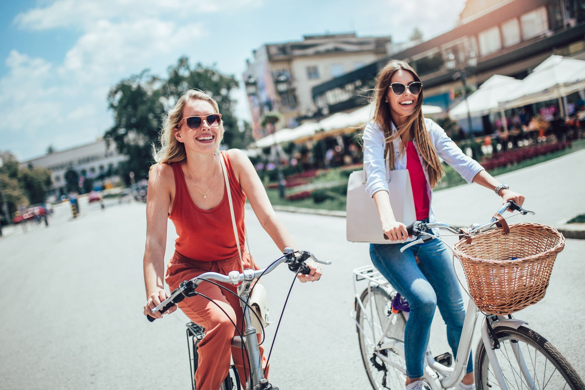 Two women on bicycles, smiling. Sunny day, city street. One wears red, the other blue jeans; both have sunglasses.