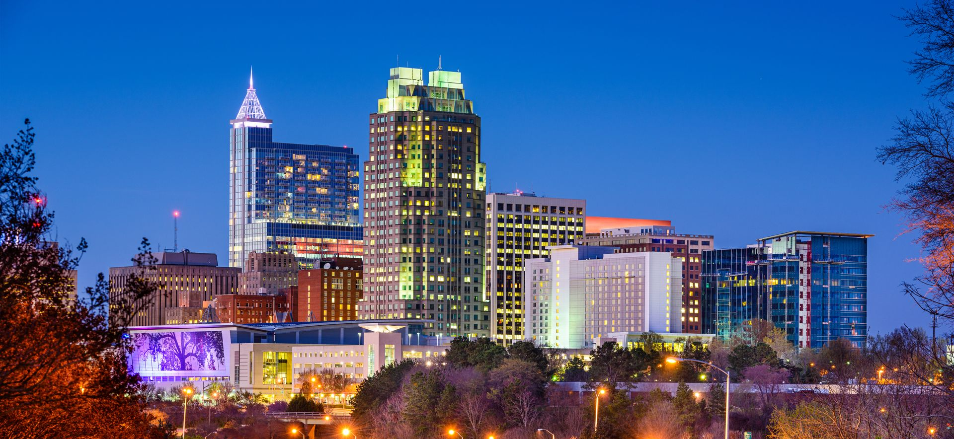 Skyline of Raleigh, North Carolina at dusk with brightly lit buildings against a blue sky.