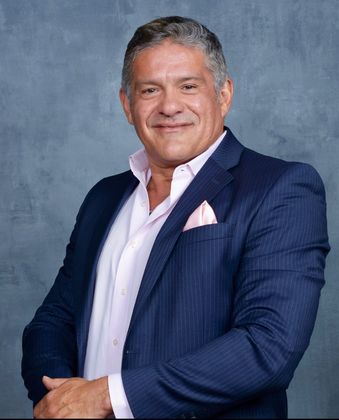 Man in blue suit with open white shirt, pink pocket square, smiling against a gray backdrop.