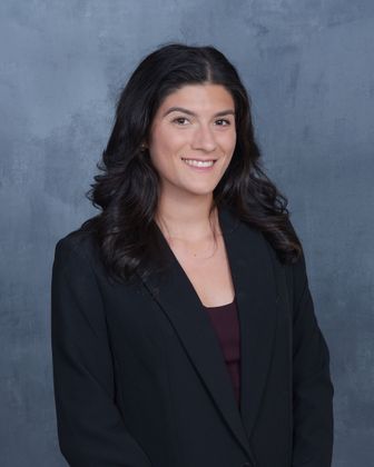 Woman with dark hair in a black blazer smiles, posing in front of a gray-blue background.