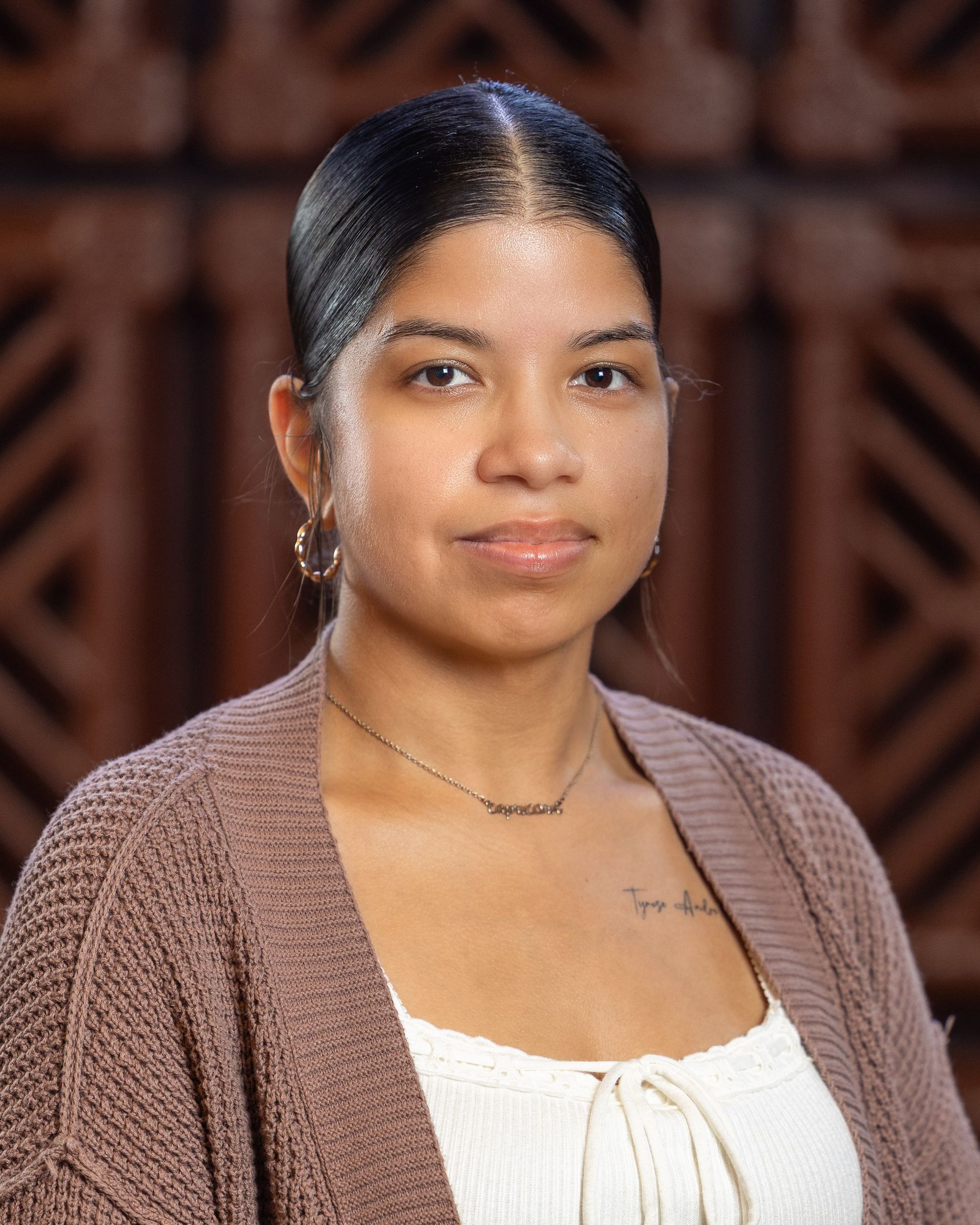 Woman wearing a white top and brown cardigan, small necklace, and earrings, against a carved wooden background.