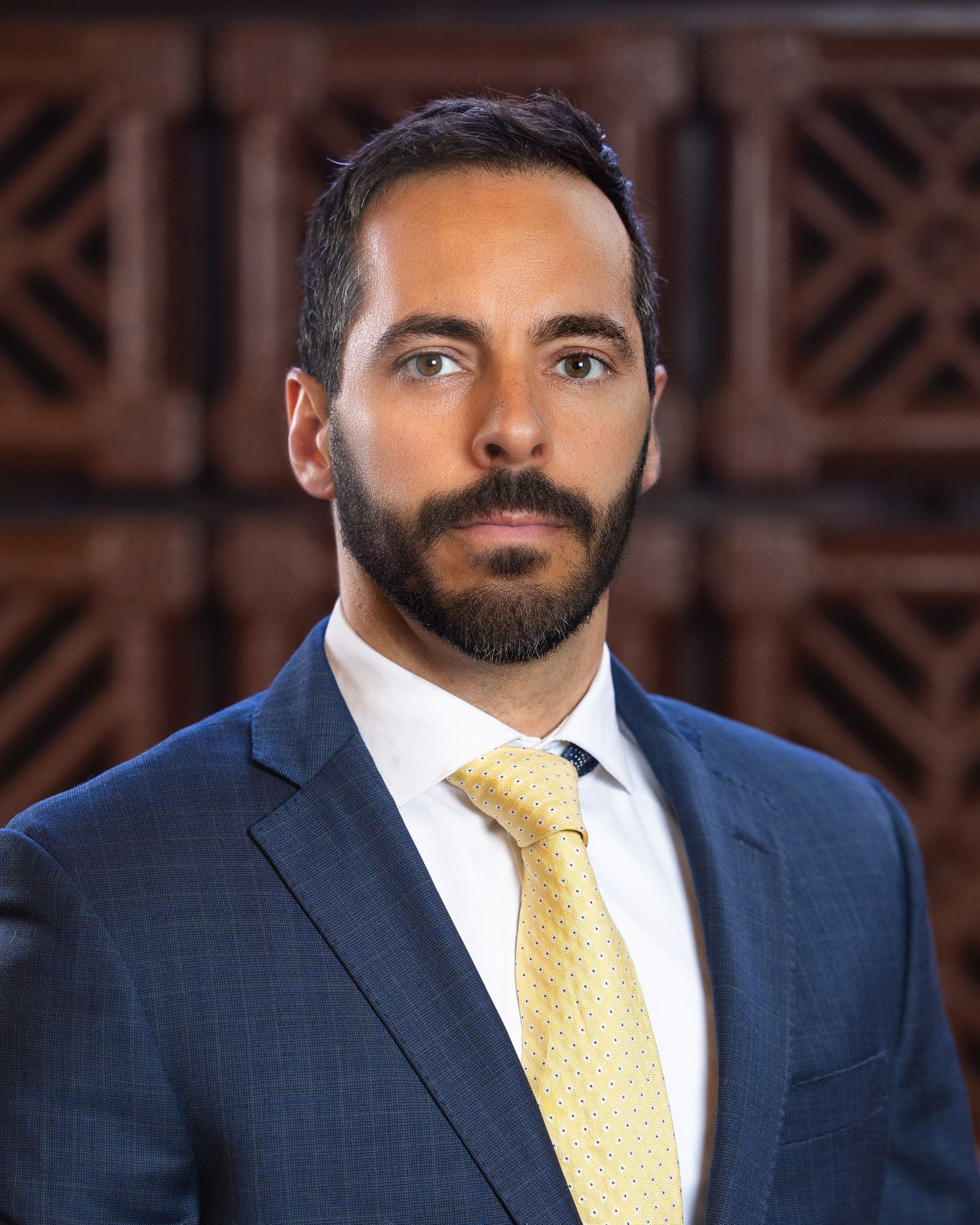 Man in a blue suit with yellow tie, serious expression, in front of a wood-paneled backdrop.