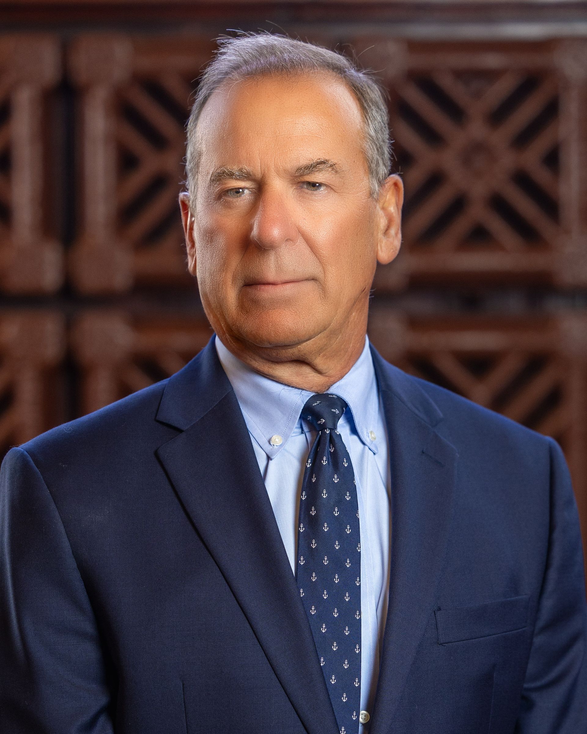 Man in a blue suit and tie, smiling slightly, standing in front of a wooden backdrop.