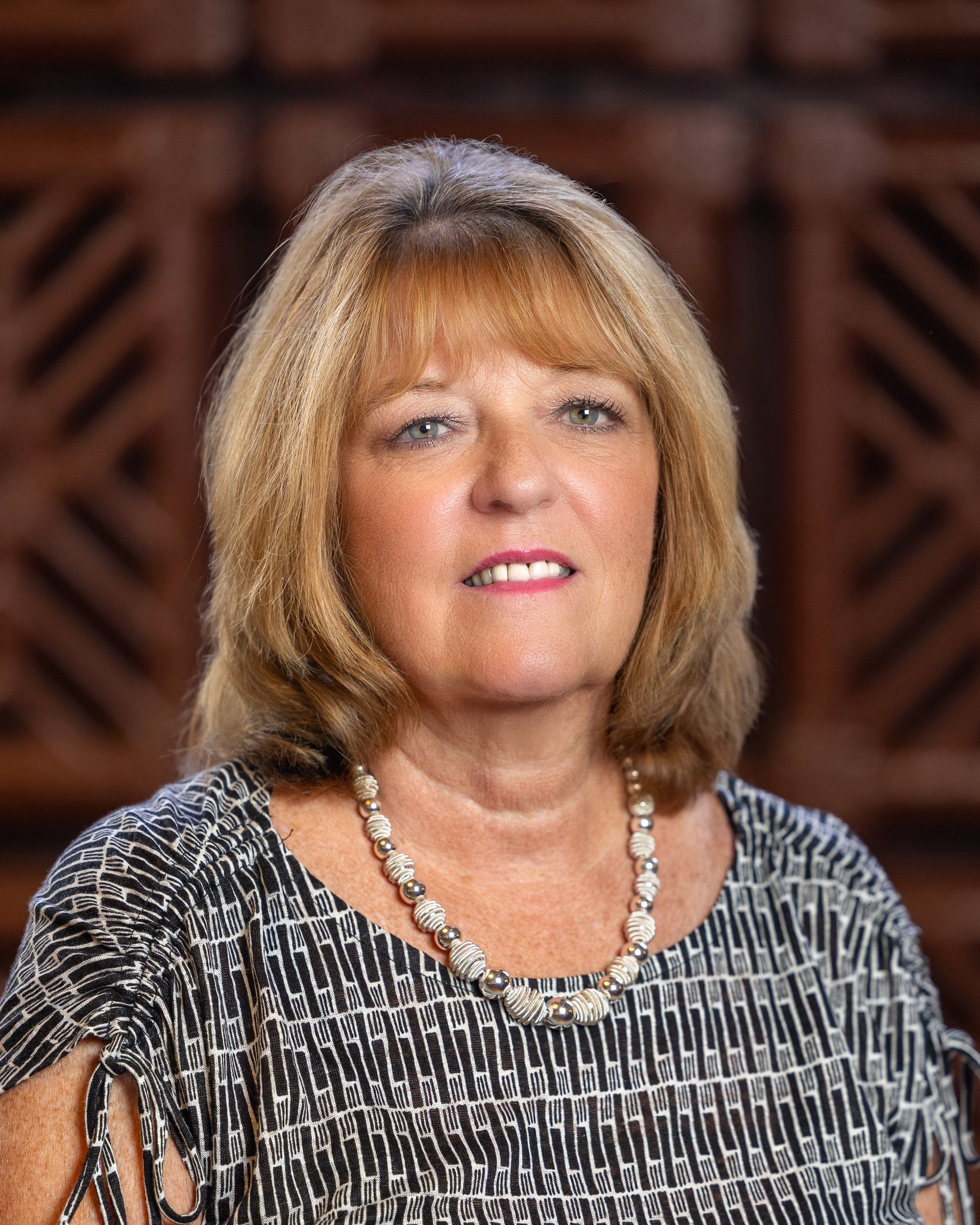 Woman with blonde hair wearing a patterned top and necklace, set against a wood background.
