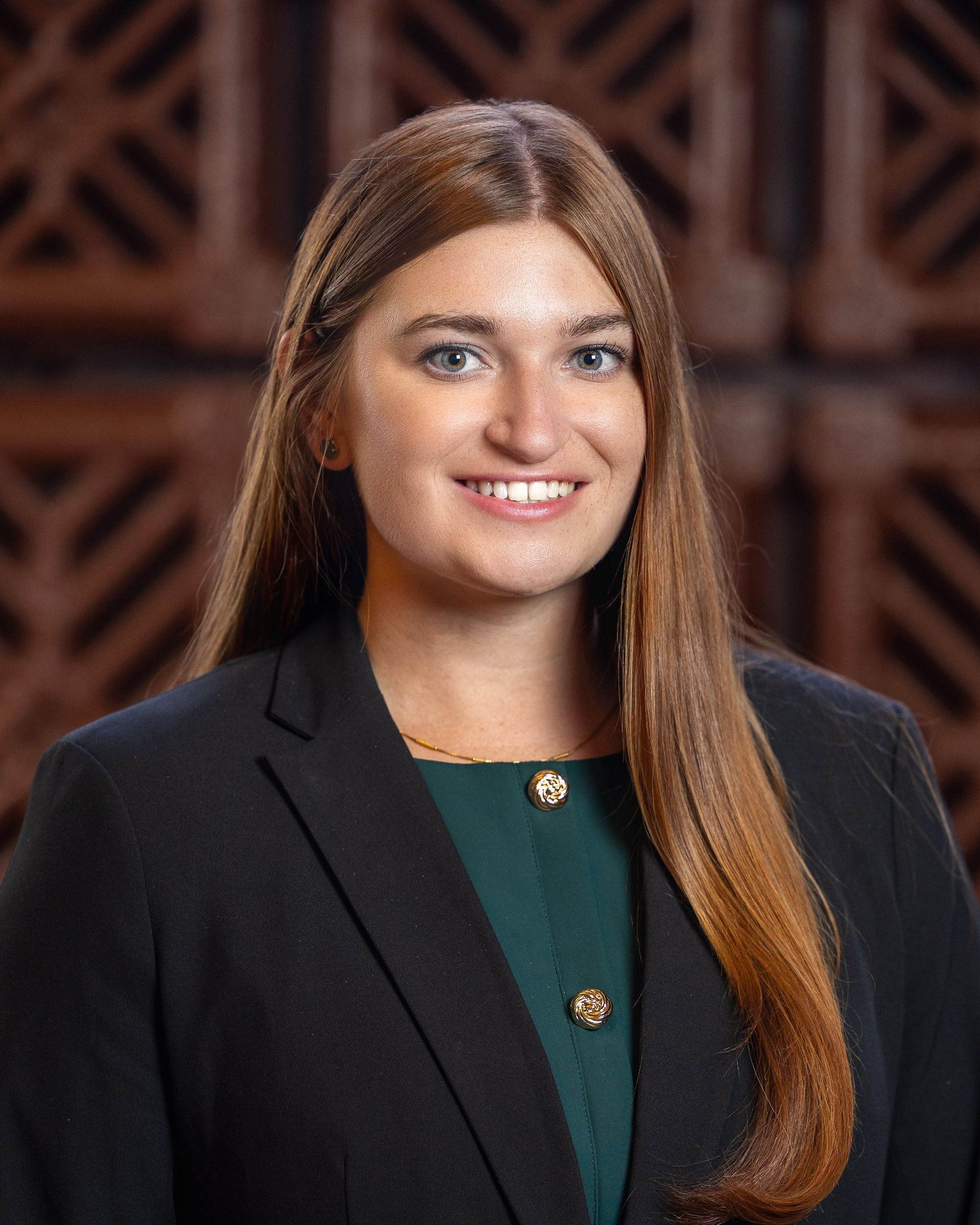 Woman in business attire smiles at the camera, dark blazer over a green top, brown hair.