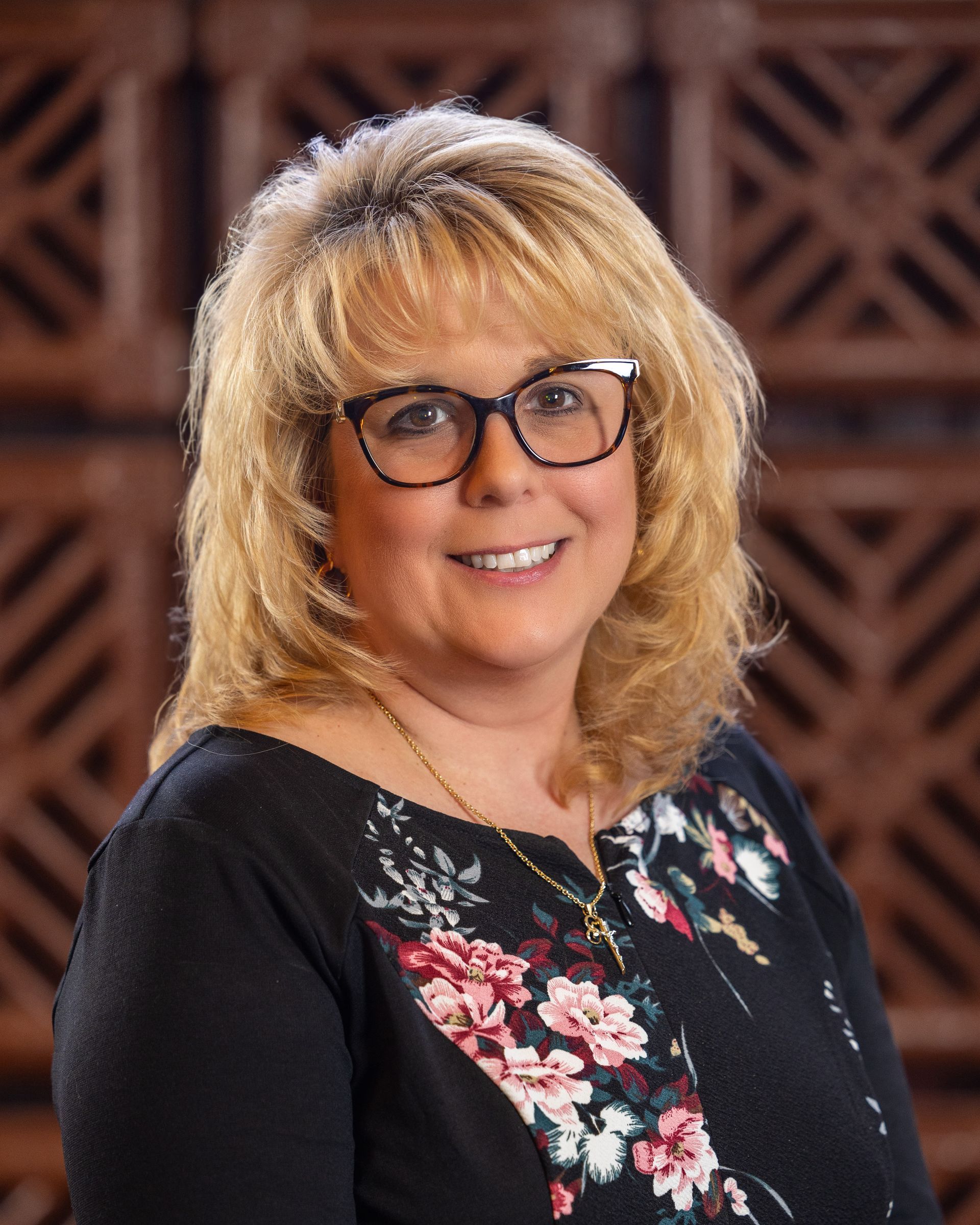 Woman with blonde hair and glasses smiles. Black floral top; brick background.