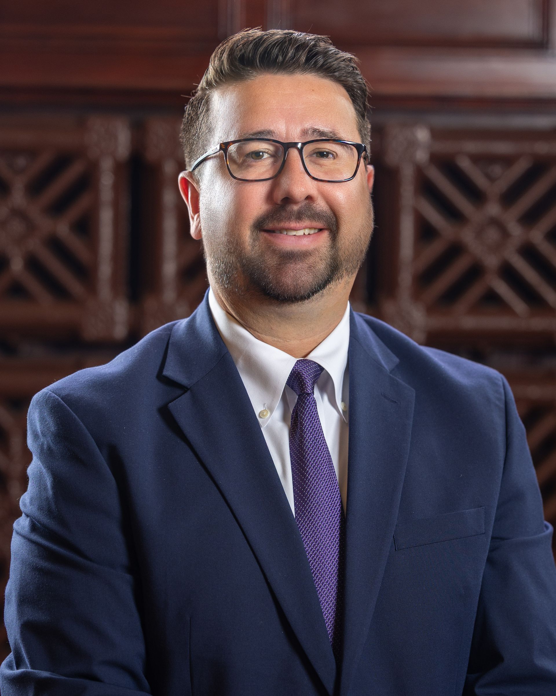 Man wearing glasses and suit, smiling in front of a wood paneled wall.