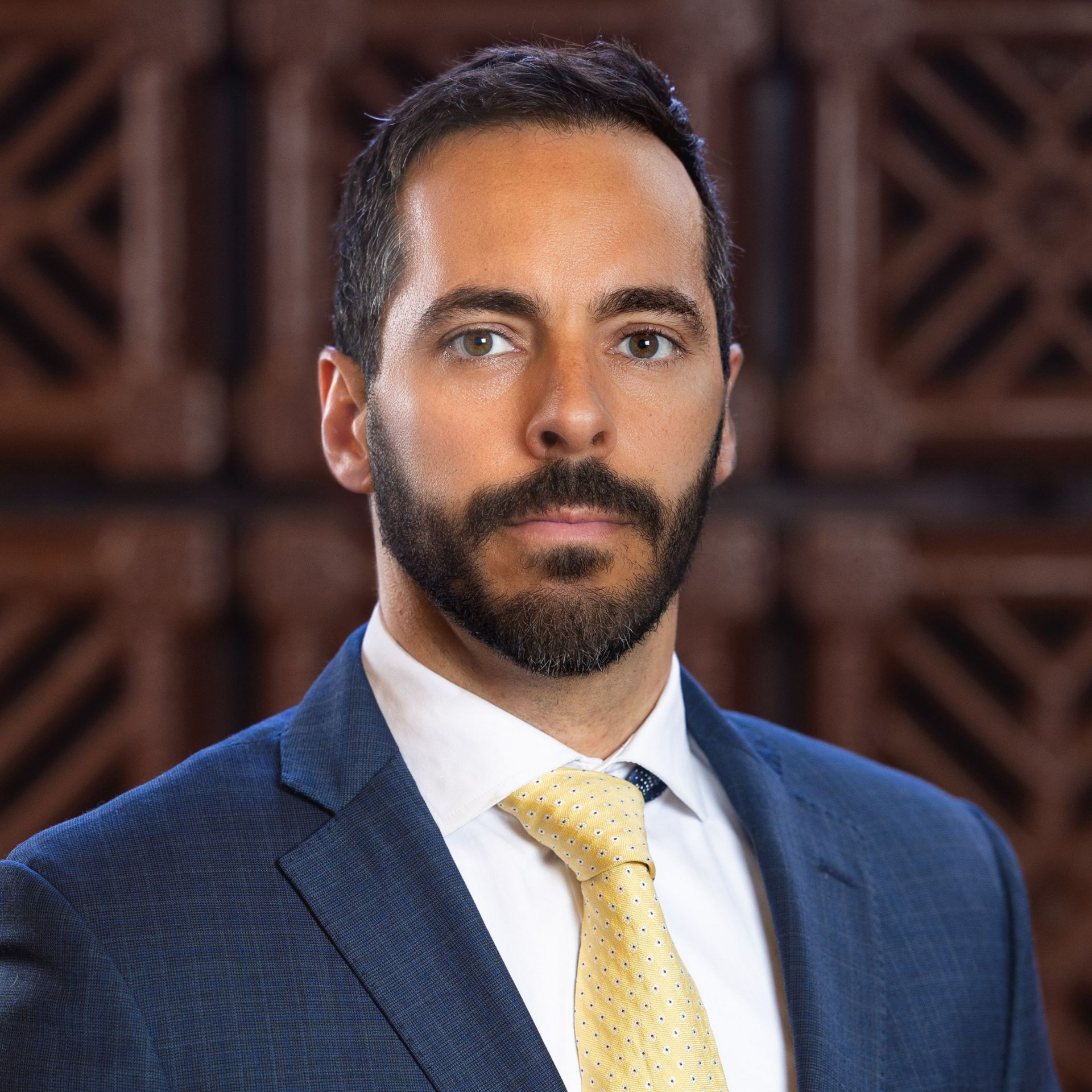 Man with dark hair and beard, wearing a blue suit and yellow tie, in front of a wooden background.