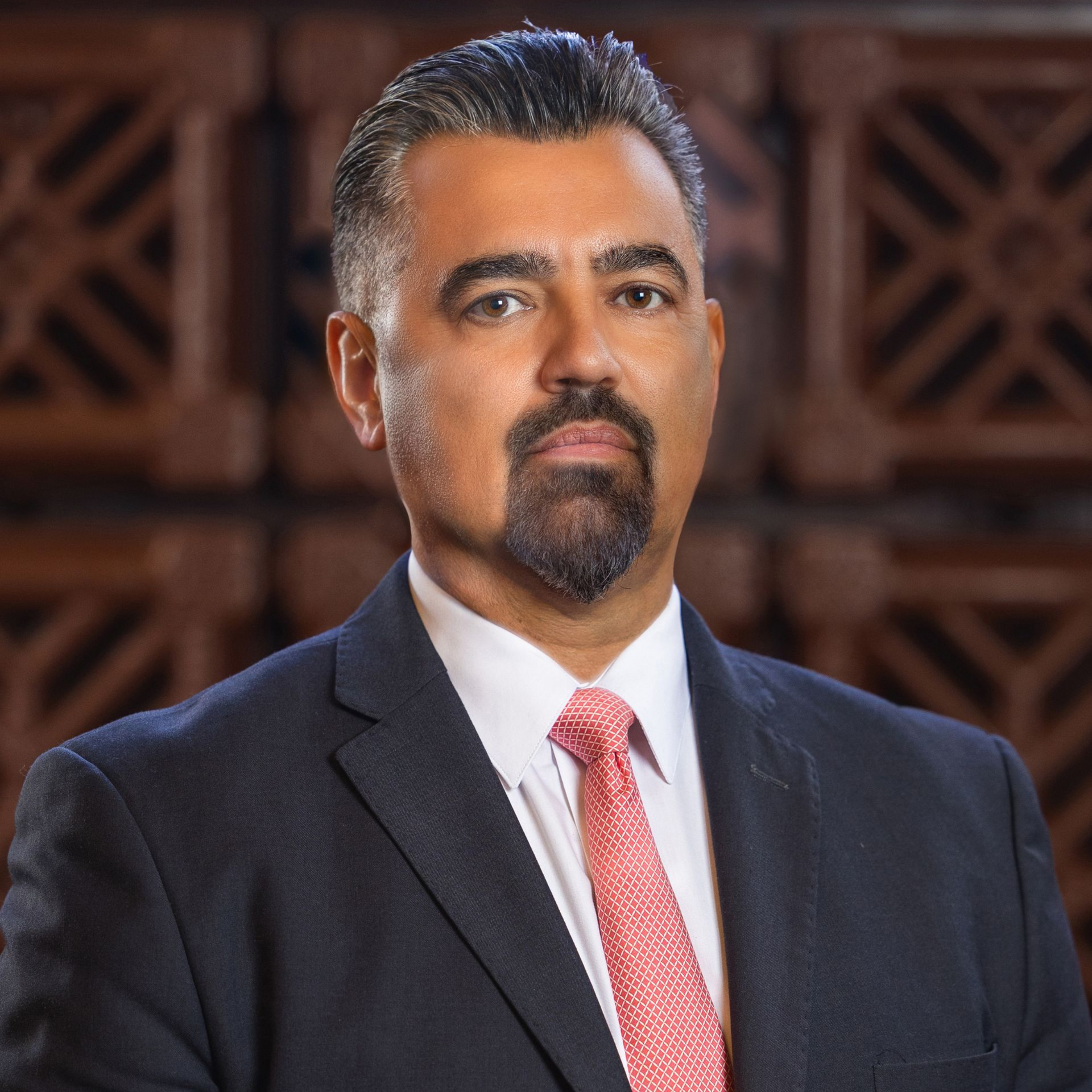 Man in suit and tie, against wood background, looking directly at the viewer.