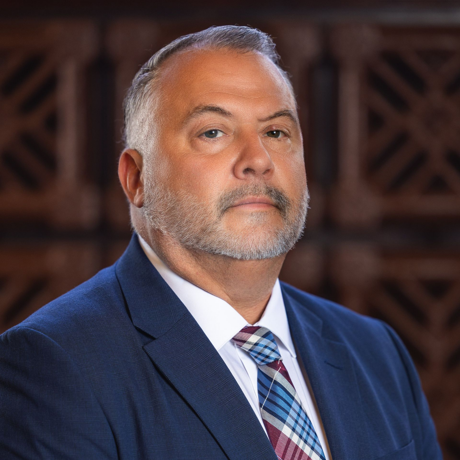 Man in a blue suit and patterned tie, standing in front of a wood panel background.