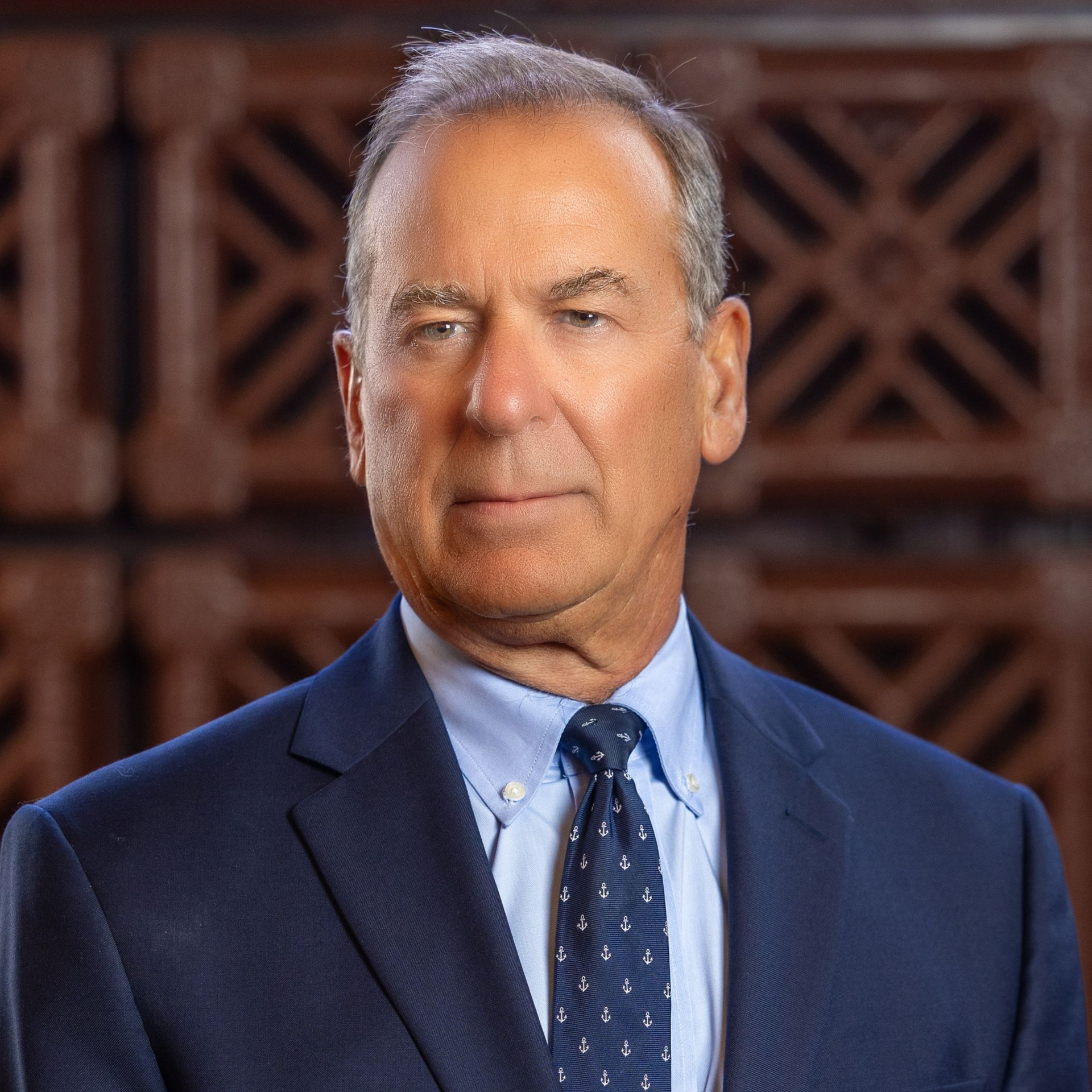 Man in blue suit, light blue shirt, patterned tie, looking forward, neutral expression, indoors.