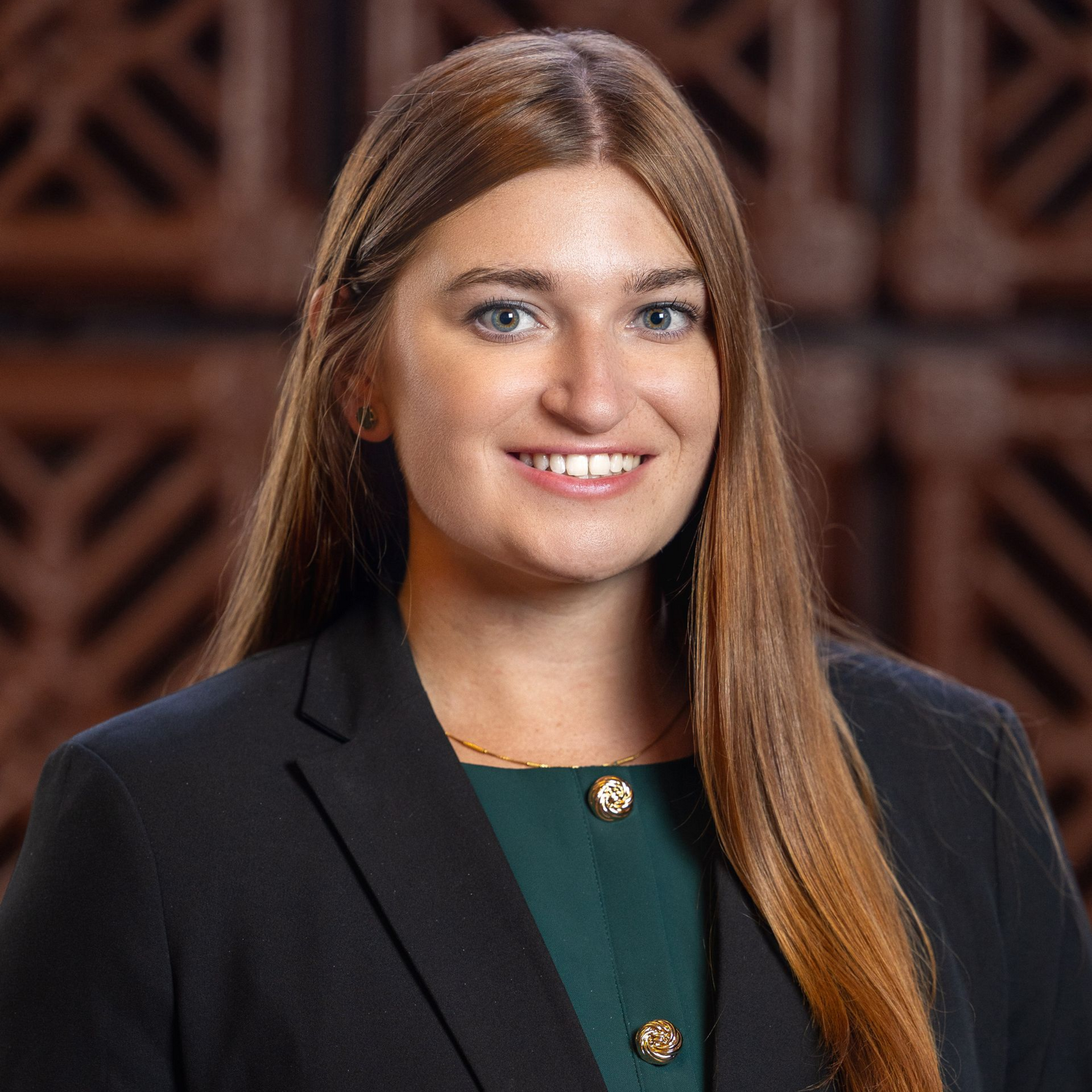 Woman with long brown hair, wearing a black blazer over a green top, smiling. Background with architectural details.