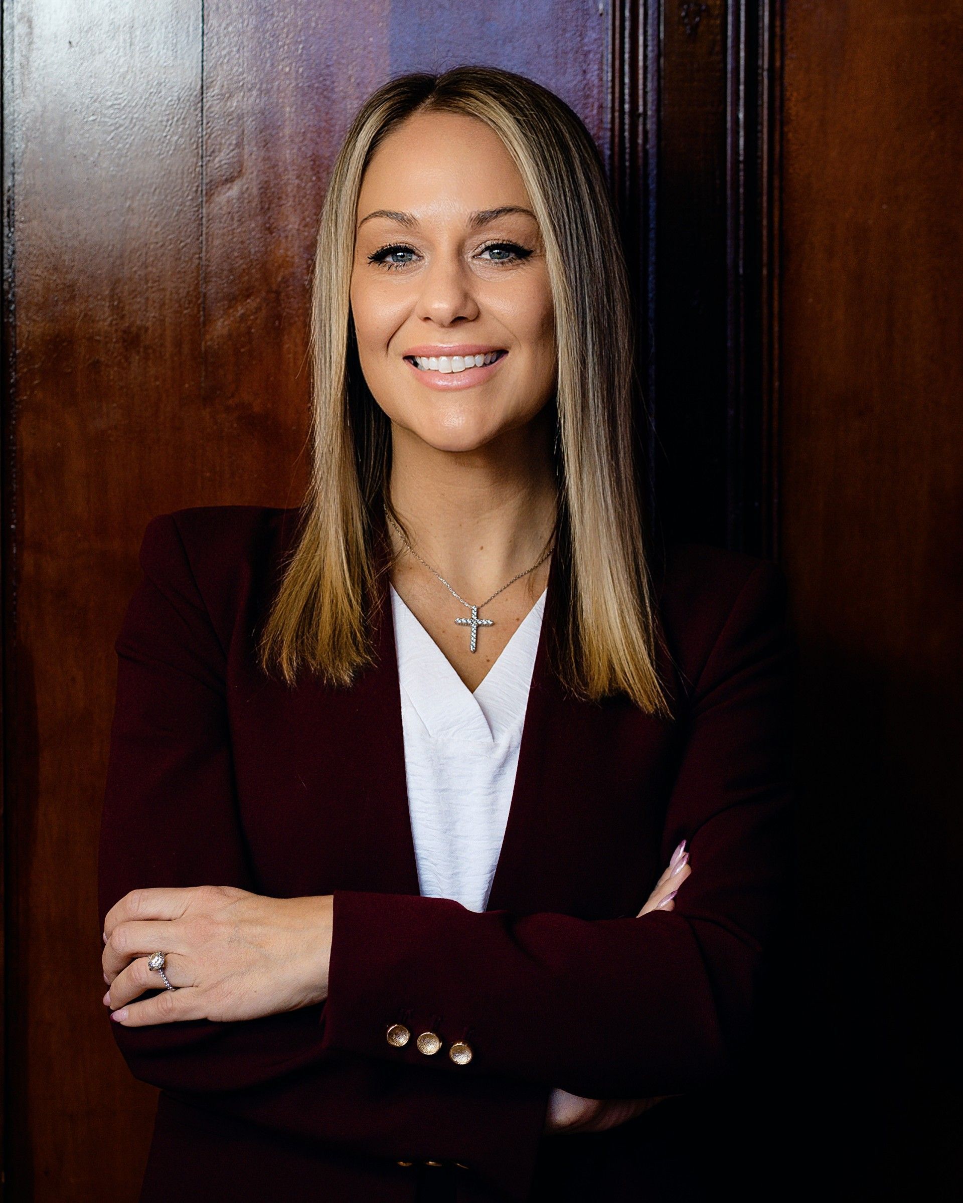 Woman in maroon blazer smiles, arms crossed, in front of a wood door.