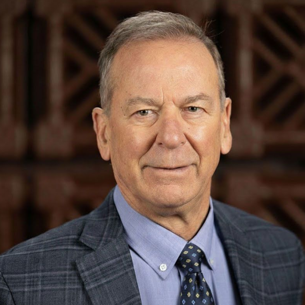 Man in blue suit, light blue shirt, patterned tie, looking forward, neutral expression, indoors.
