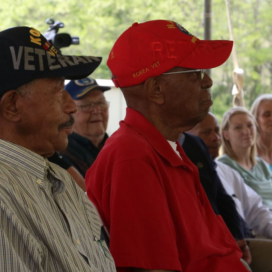 Two veterans in hats at an outdoor event. One wears red, the other a navy hat. People sit in the background.