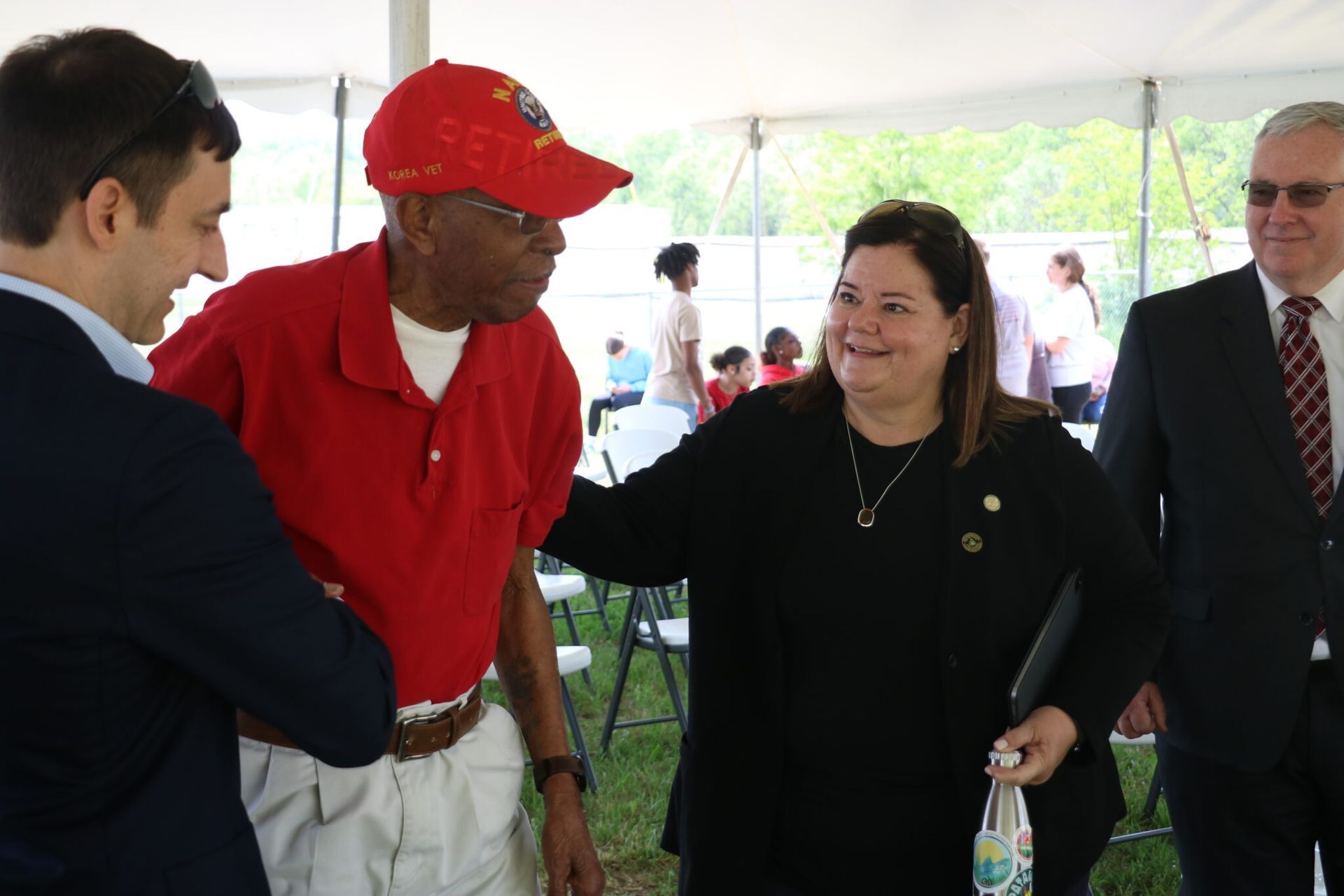 People interacting outdoors; man in red shirt and hat smiles as others greet him.