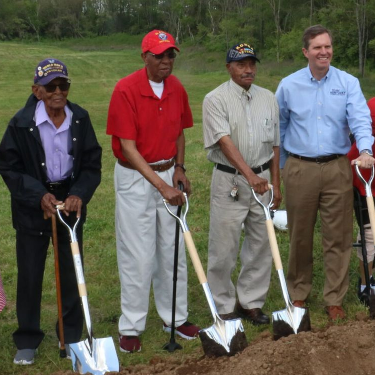 Men with shovels at a groundbreaking ceremony, standing on dirt. They wear casual clothes.